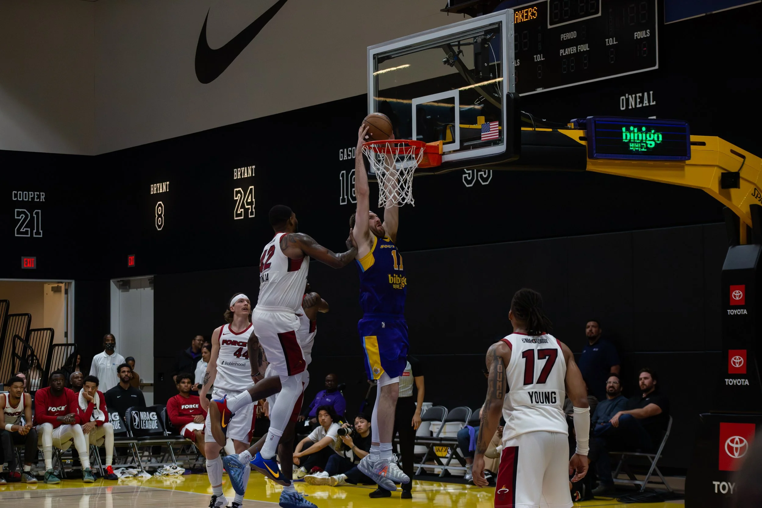 A basketball game in progress with a player in a blue and yellow jersey jumping to make a basket while an opposing player in a white and red jersey attempts to block the shot. Other players and spectators watch from the sidelines inside an indoor bas