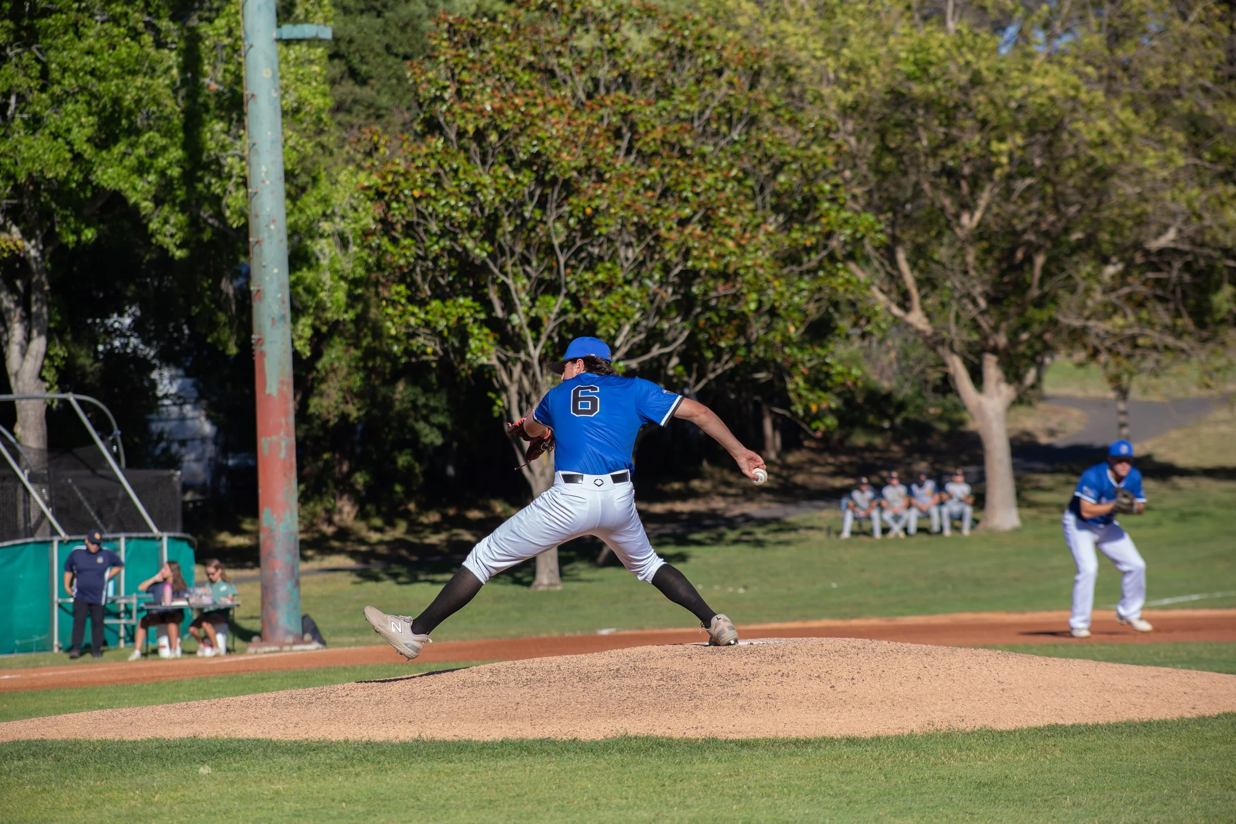 A baseball pitcher in a blue jersey with the number 6 throws a pitch on a baseball field. The pitcher is mid-motion, with the team and spectators visible in the background, and tall trees lining the field.