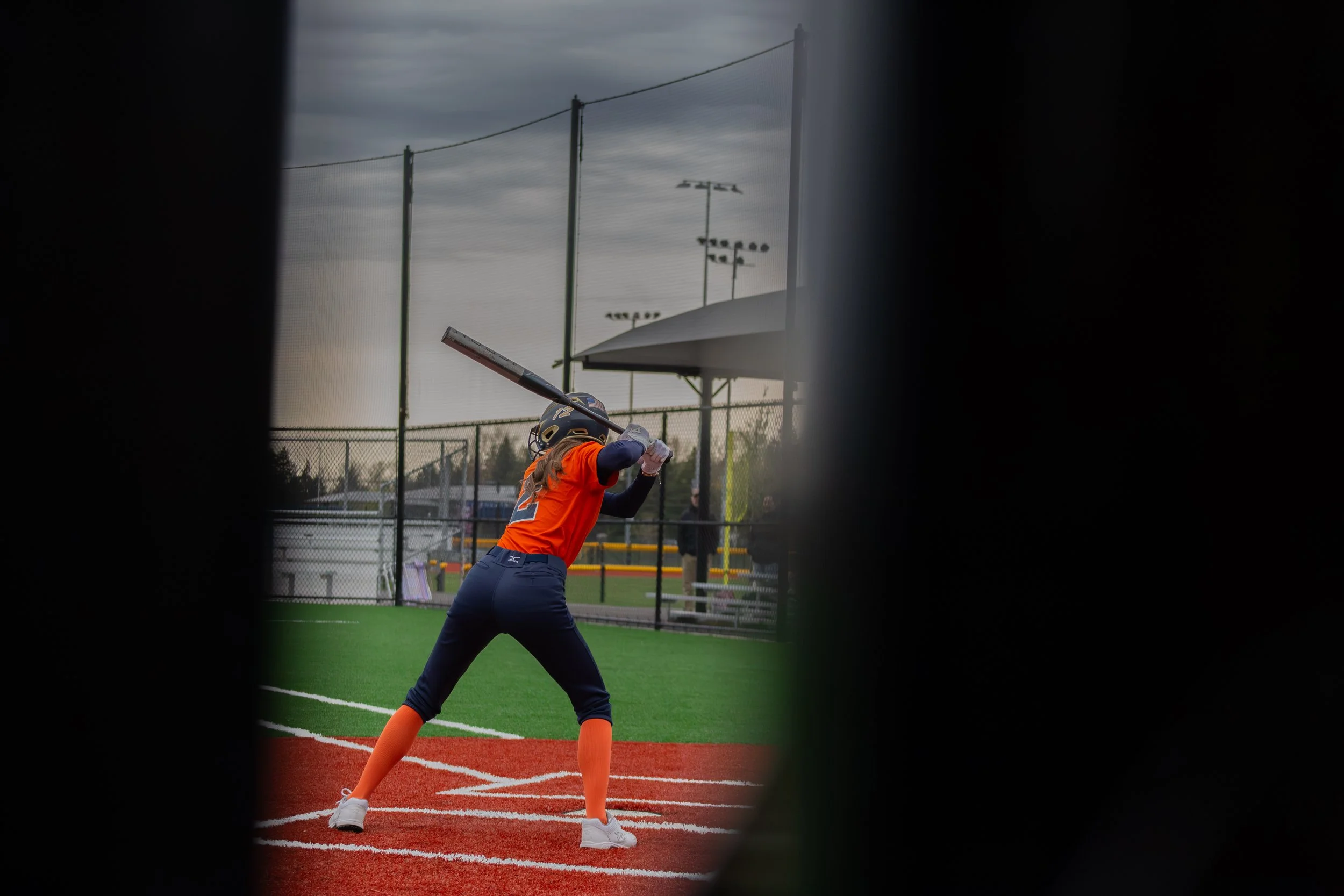 A female baseball player in an orange jersey and dark pants is swinging a bat at a baseball field, viewed through a narrow opening.