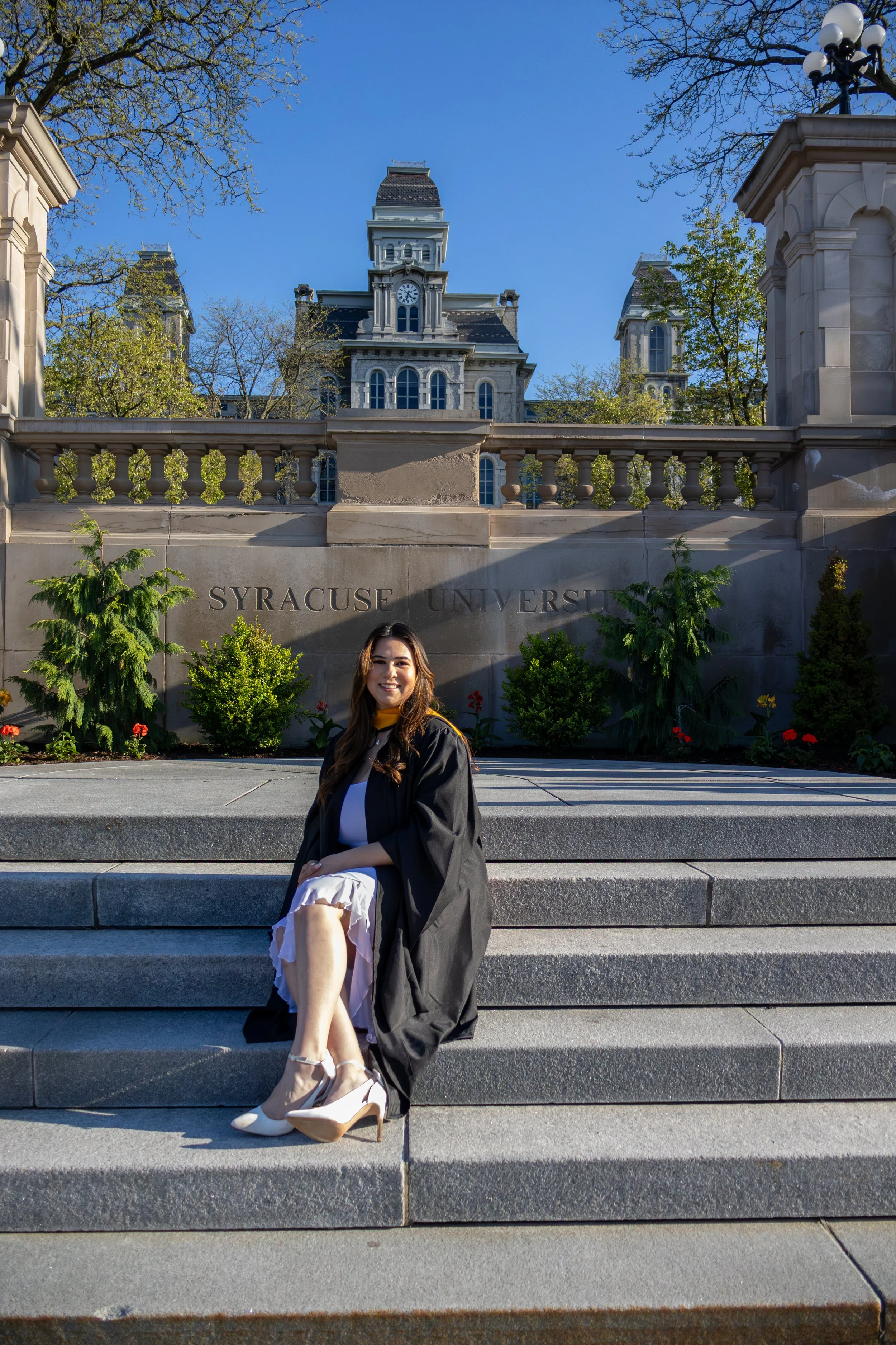 A smiling woman in graduation attire sitting on the steps in front of a university building with the sign 'Syracuse University' visible.