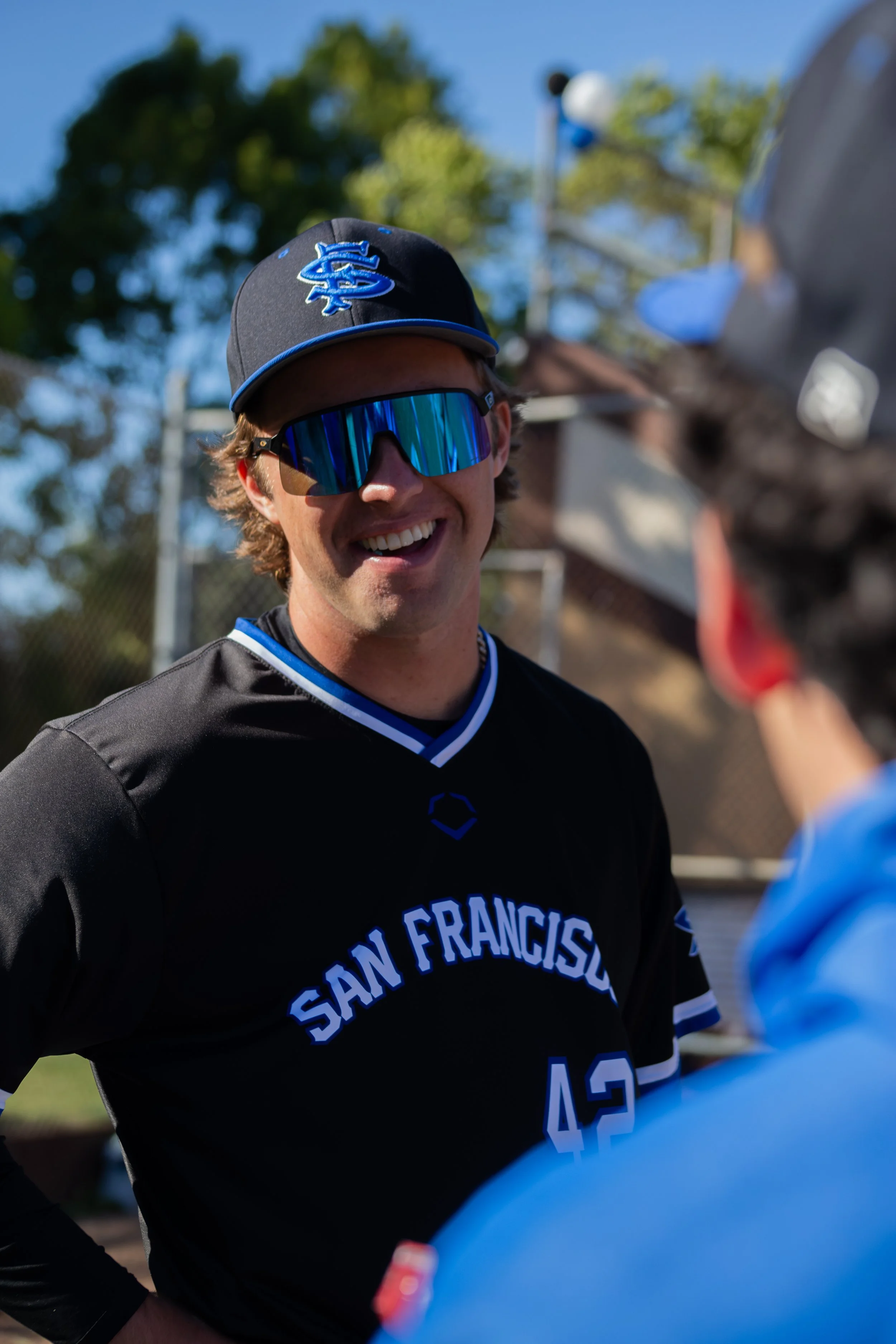 A smiling man wearing sunglasses, a baseball cap with a blue logo, and a black sports jersey with 'San Francisco' and the number 43, engaging in conversation outdoors on a sunny day.
