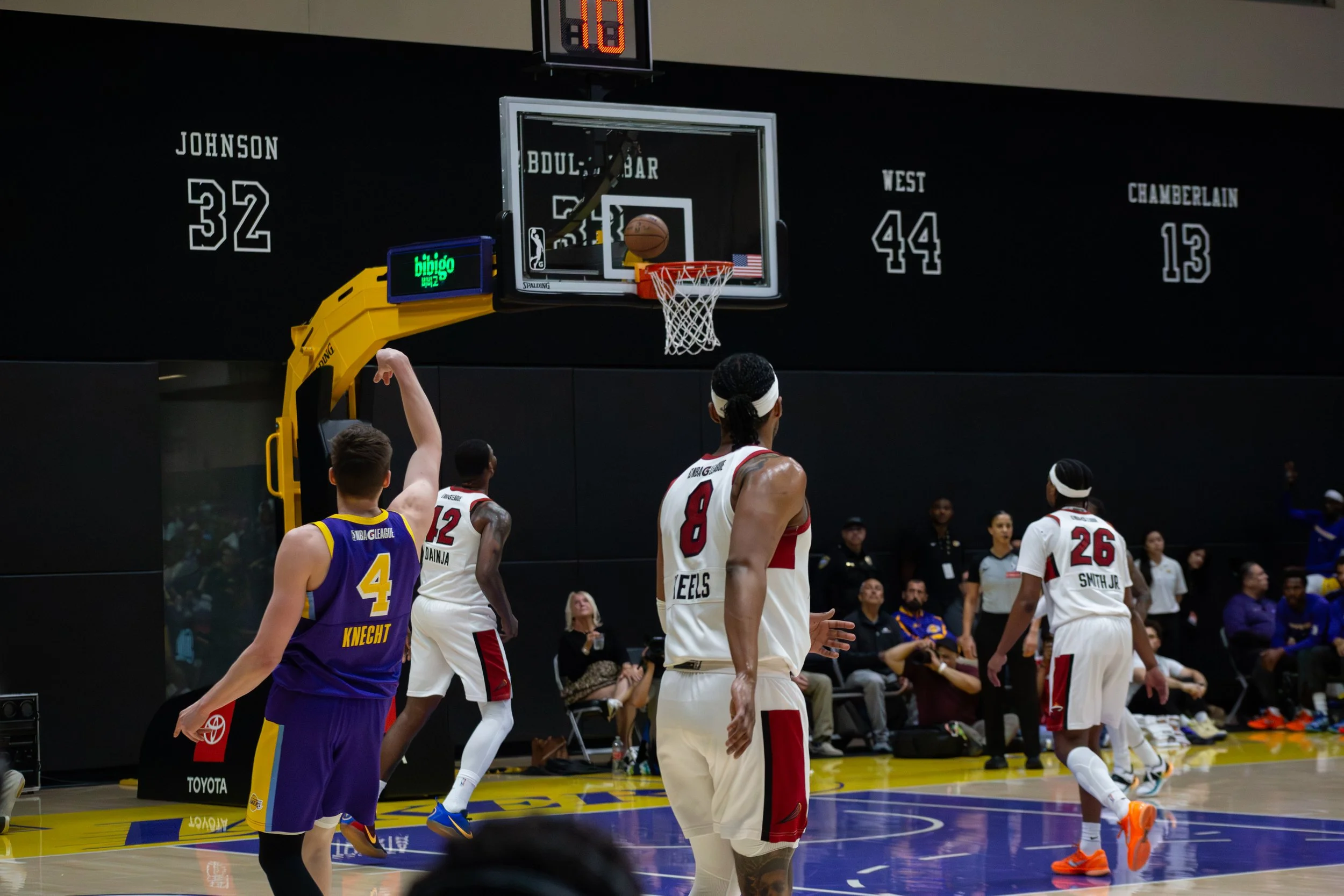 A basketball game in progress featuring players from two teams. One team is wearing white jerseys with red and black accents, and the other team is wearing purple and yellow jerseys. The scene shows several players on the court and spectators watchin