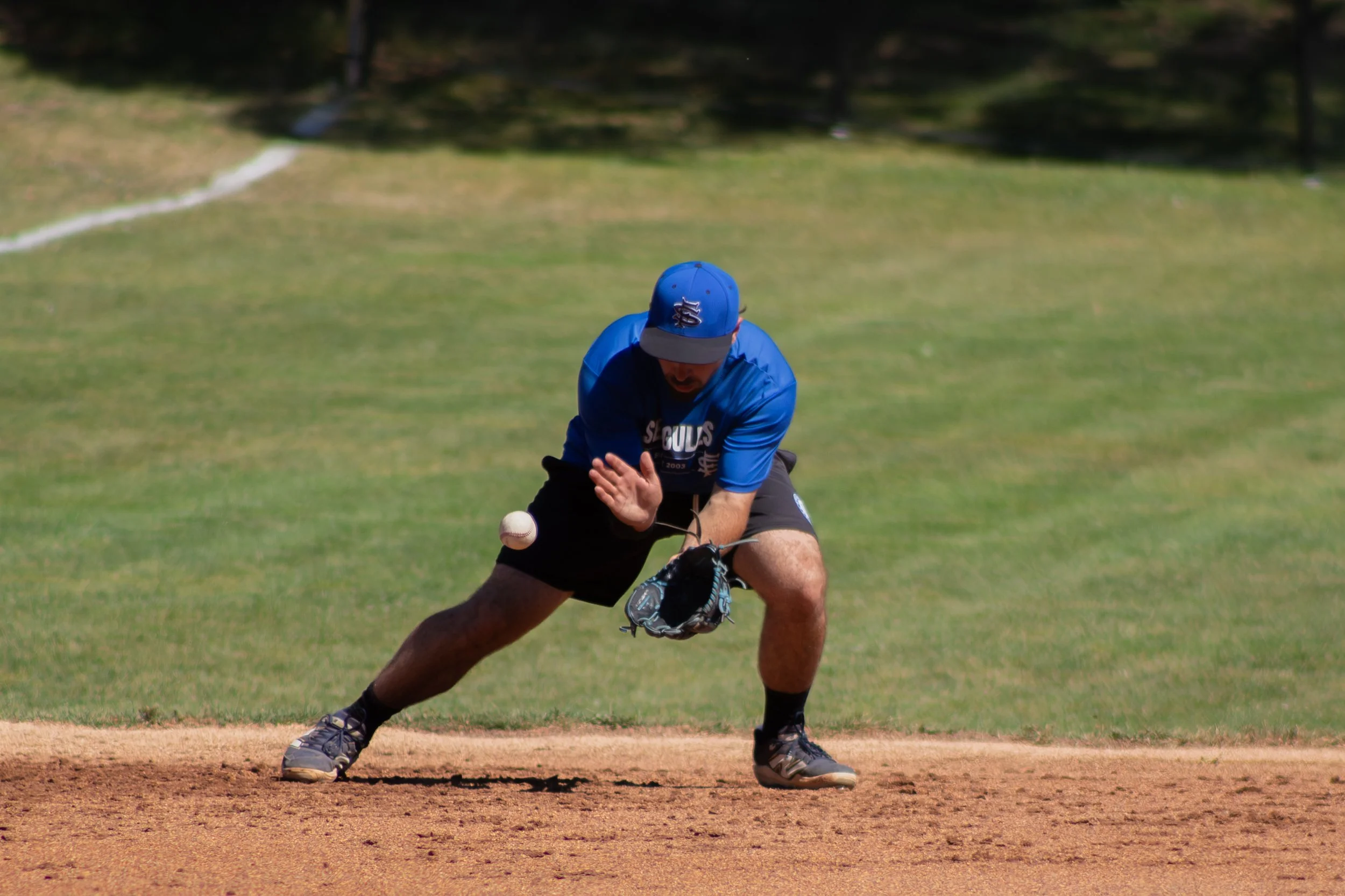 A man in a blue baseball cap and shirt playing baseball, crouched on a field with a baseball and glove.