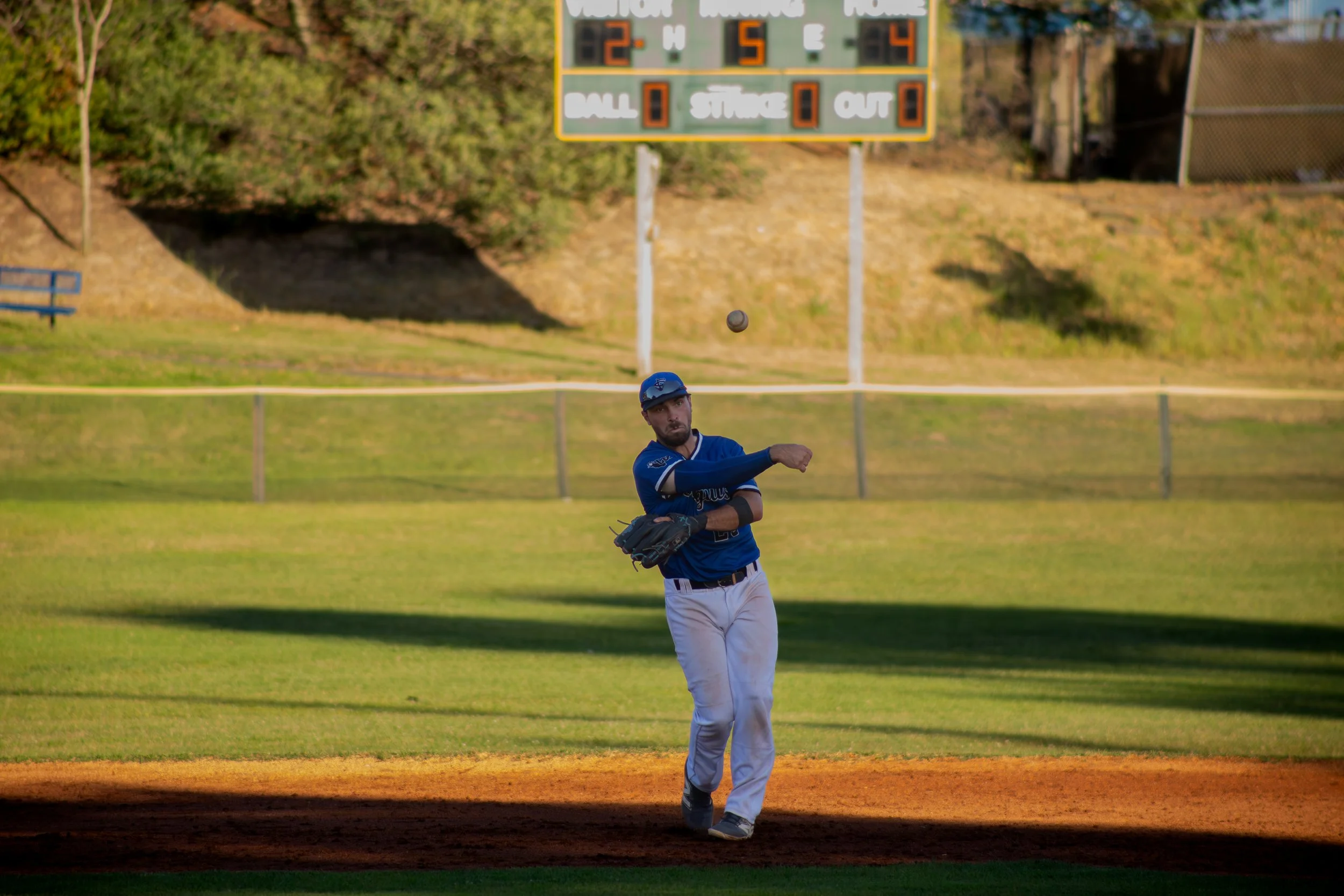 A baseball player wearing a blue jersey, white pants, and a blue cap preparing to catch a baseball on a baseball field with a scoreboard in the background.