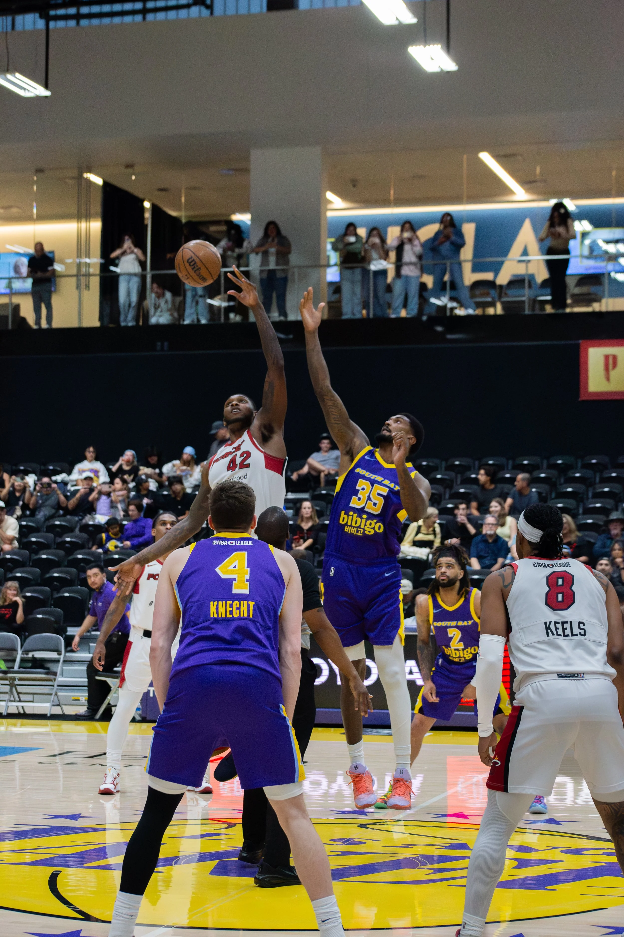 A basketball game in progress with players on the court and spectators watching from the stands. Two players are jumping for the ball, one in a white jersey and the other in a blue jersey, under ceiling lights.