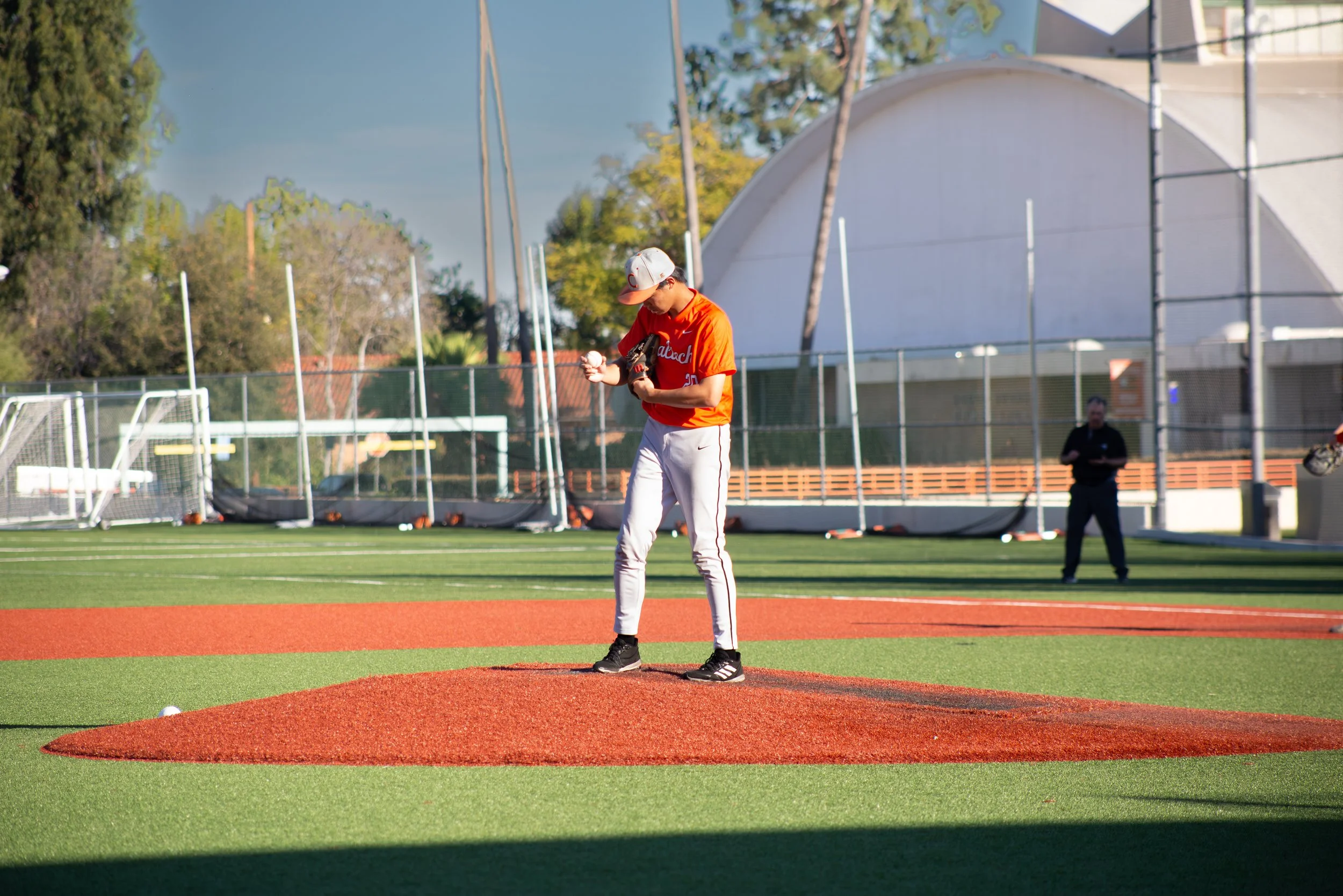 A baseball player in an orange jersey and white pants stands on the mound holding a baseball and glove, preparing to pitch on a baseball field.