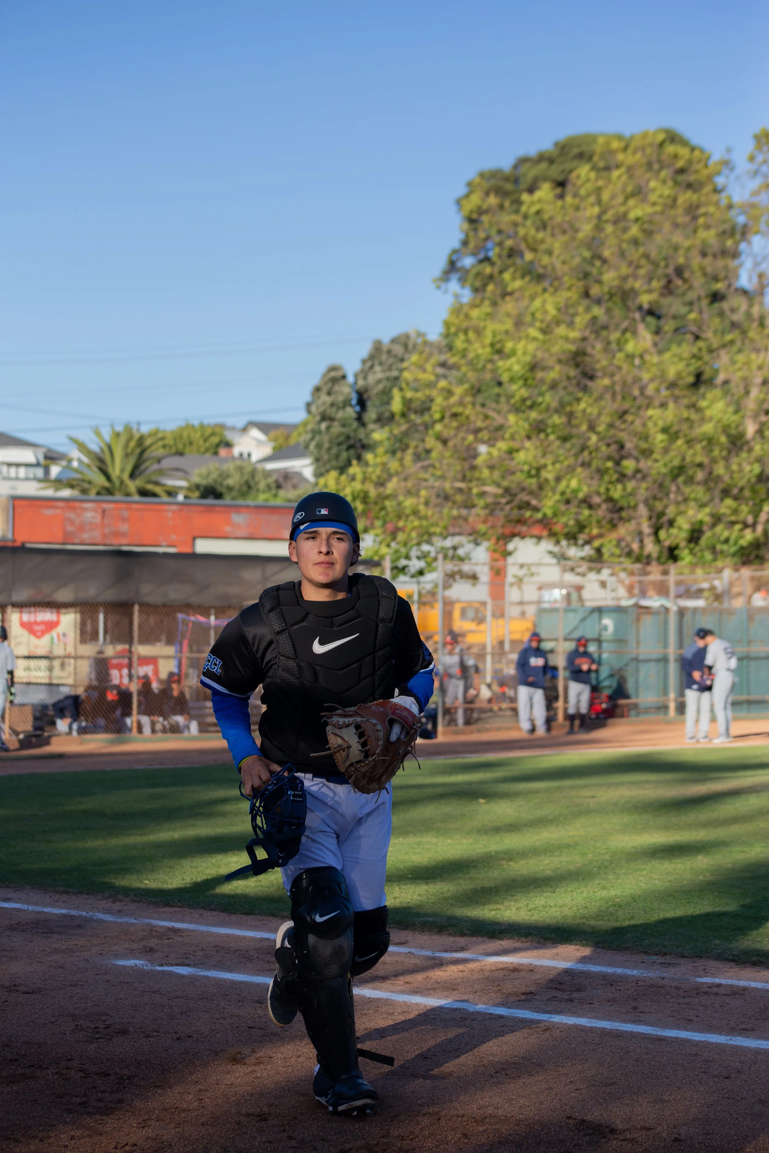 A young male baseball player in full gear running on the field, with a background of trees, a fence, and spectators.
