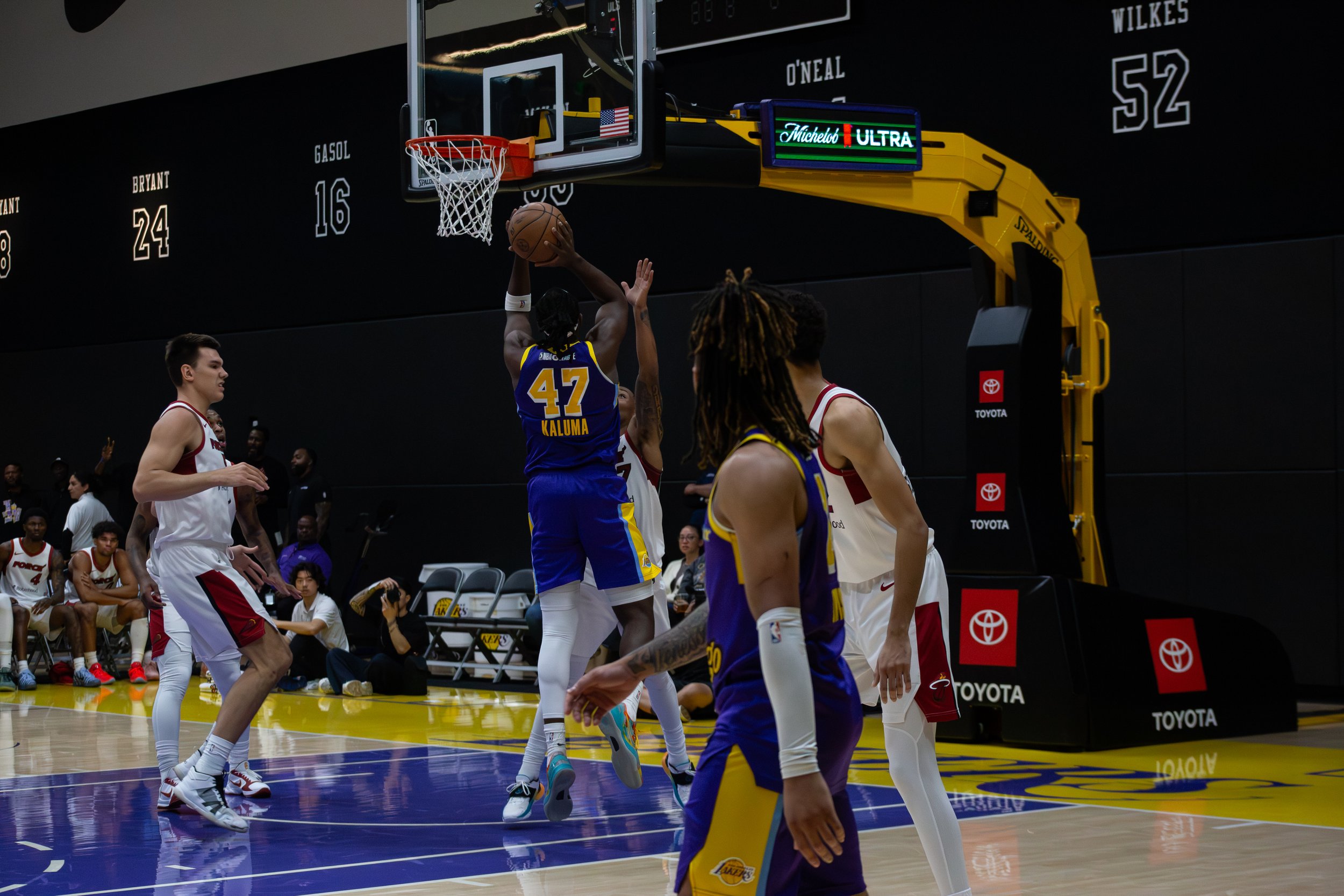 A basketball player in a blue and yellow uniform jumping to shoot the ball towards the hoop while opposing players in white and red uniforms attempt to block.