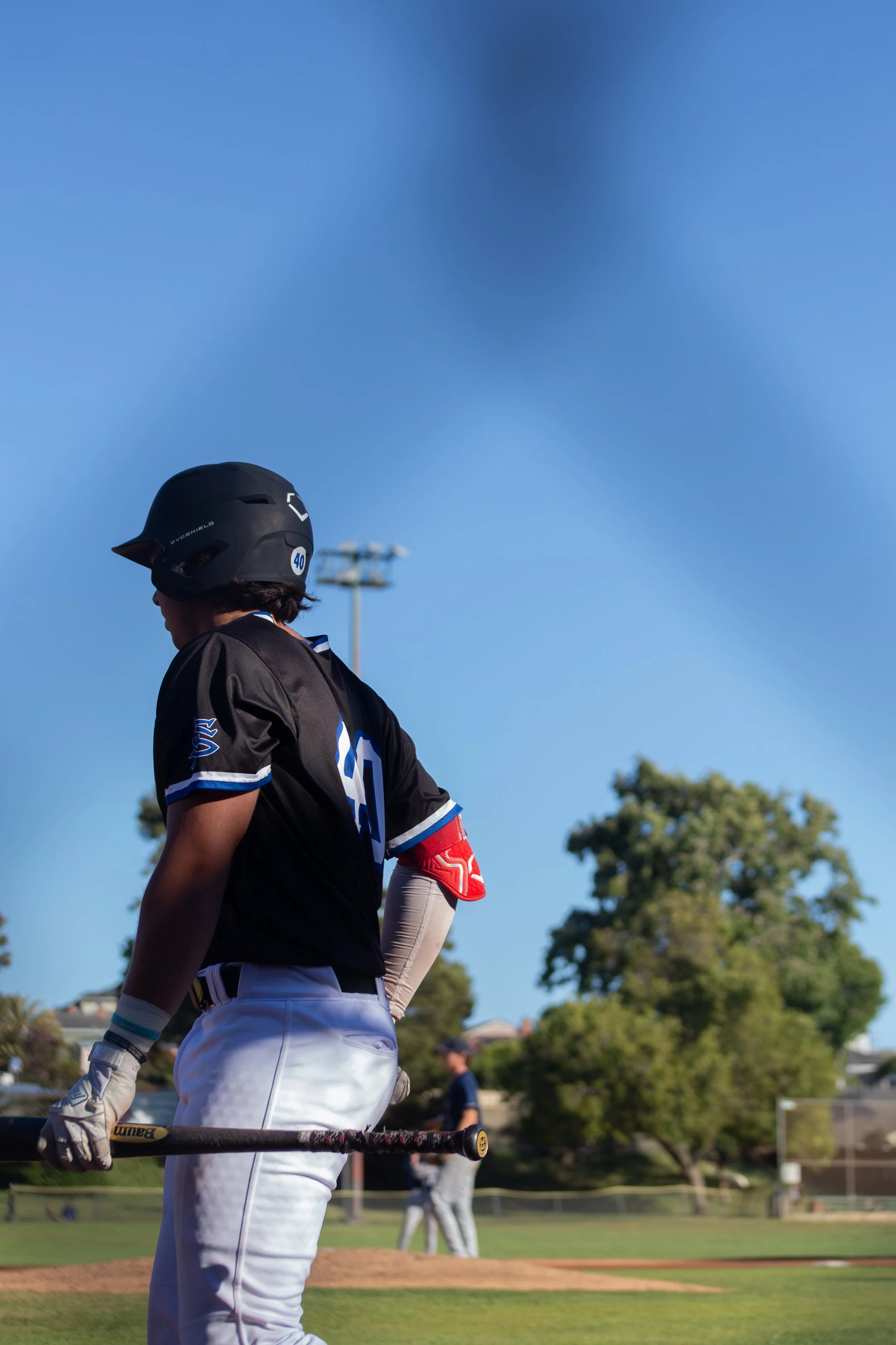 A baseball player wearing a black helmet, black jersey with blue and white accents, and white pants, holding a baseball bat, standing on a baseball field with trees and a few people in the background under a clear blue sky.