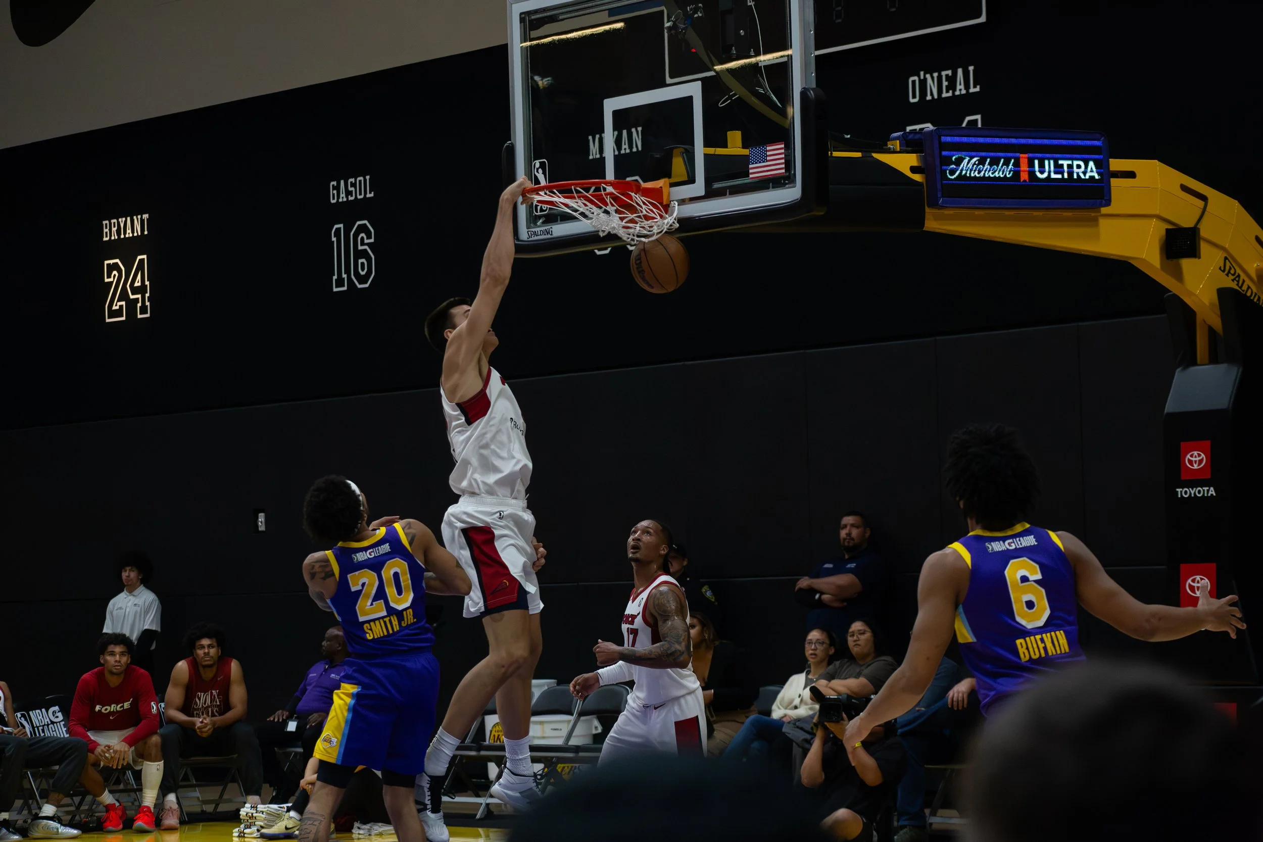 A basketball game in progress with a player dunking the ball into the hoop while other players and spectators observe. The player dunking wears a white jersey, and other players are in blue and yellow jerseys. The background includes a black wall wit
