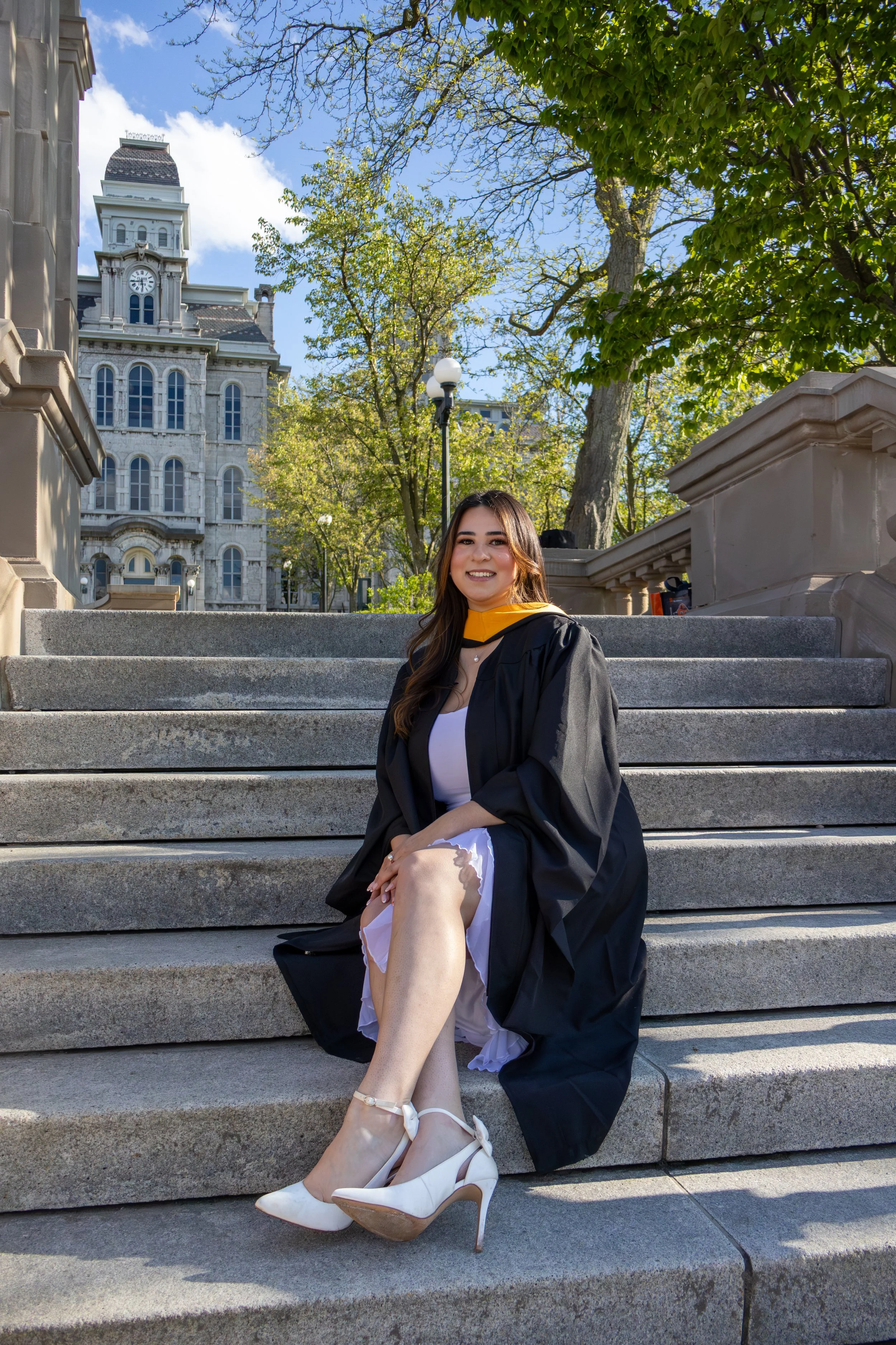 A young woman in a graduation gown seated on an outdoor stone staircase smiling with her legs crossed. She is wearing white high heels and a white dress underneath her gown. In the background, there are historic buildings, trees with green leaves, an
