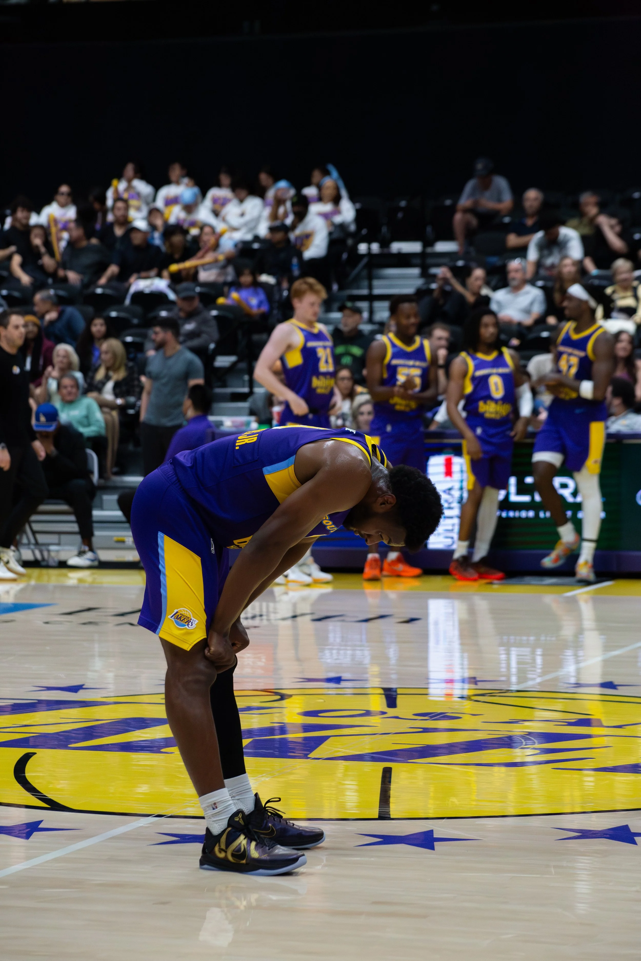 A basketball player in a blue and yellow Los Angeles Lakers uniform is bent over, with his hands on his knees, on the basketball court. In the background, other players and spectators are visible, some standing and some sitting, with a dark backgroun