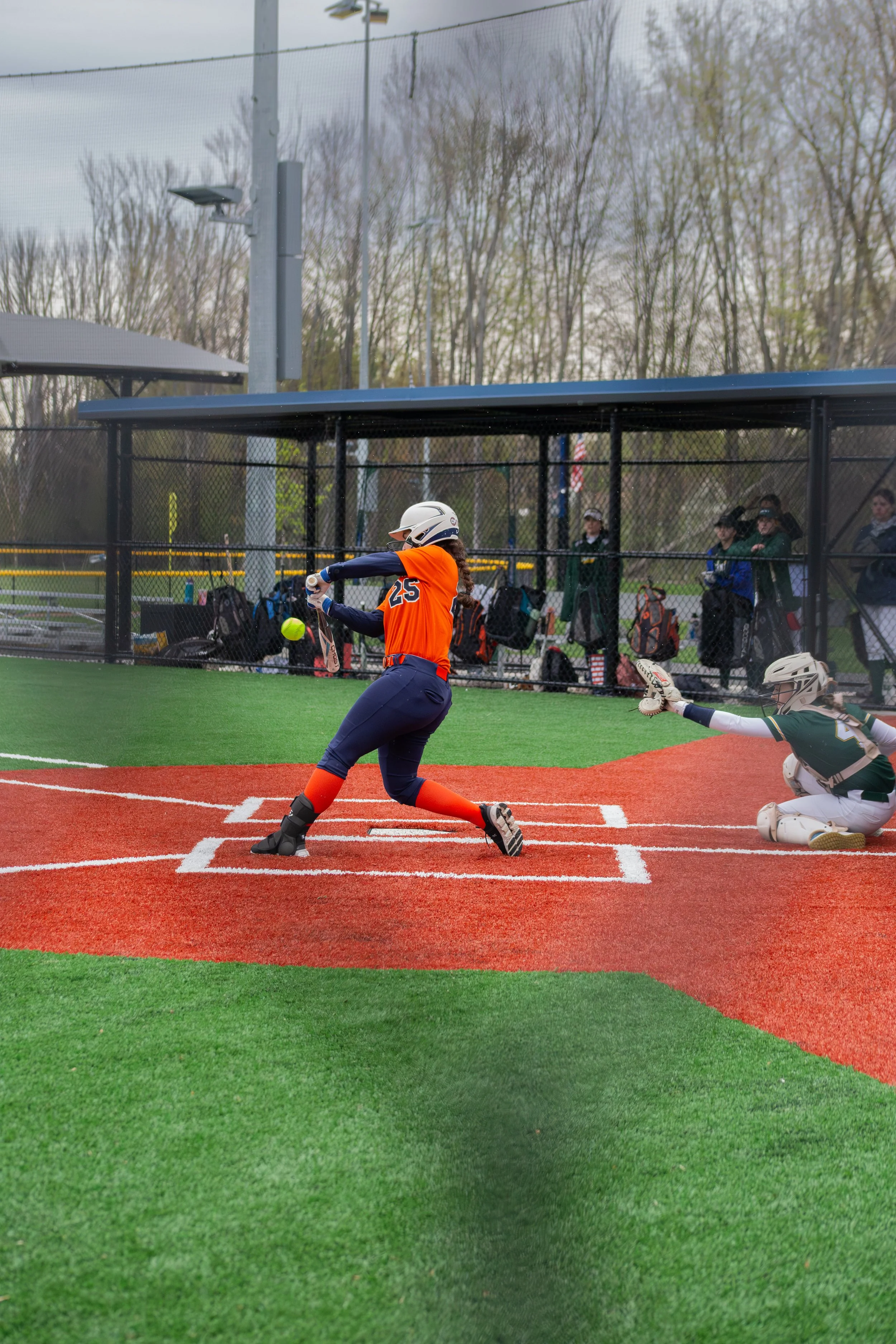 A girl in a baseball uniform, wearing an orange jersey with the number 25, is swinging a bat at a baseball in a batting cage. There is a catcher in a green and white uniform reaching out to catch the ball. Spectators are watching from behind the cage