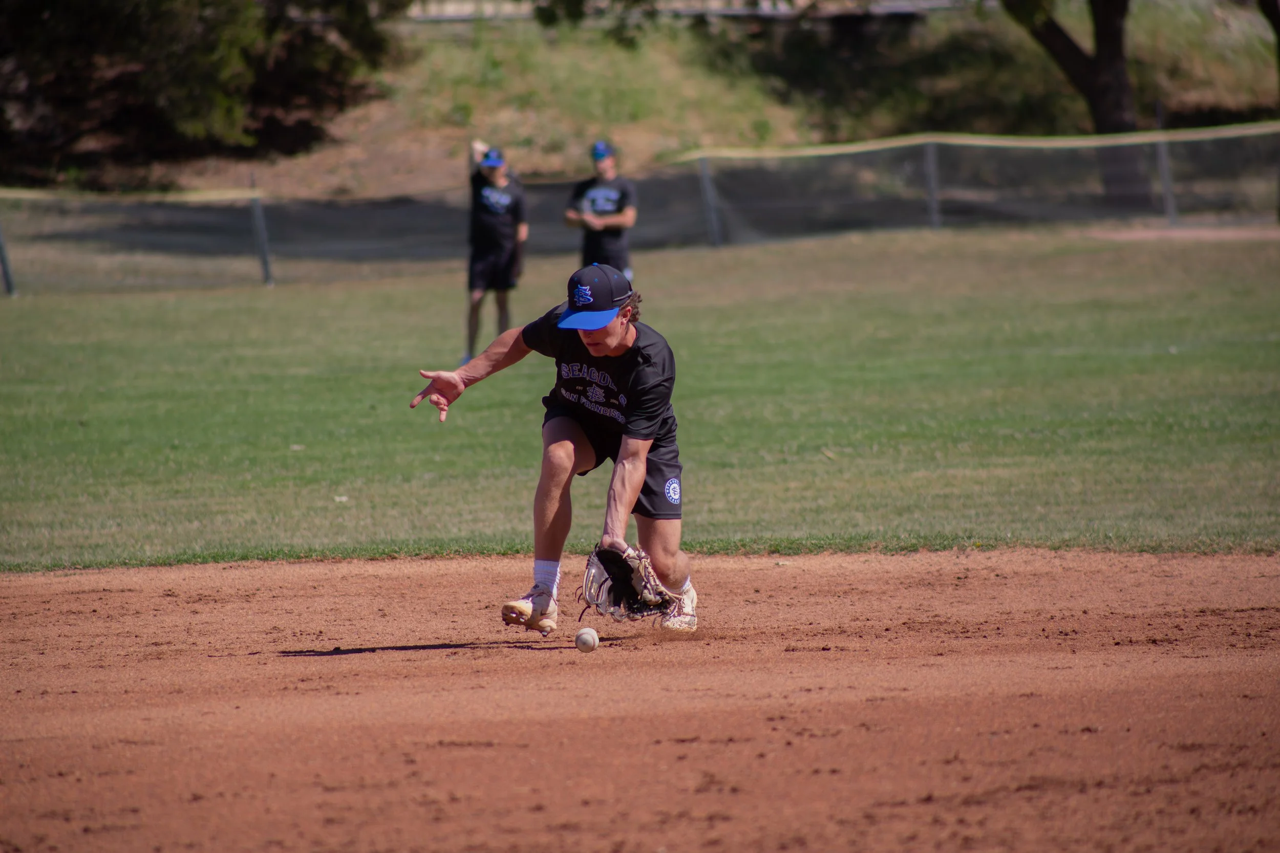 A baseball player in black and blue uniform along with a cap, extending their hand to pick up a baseball on the field.