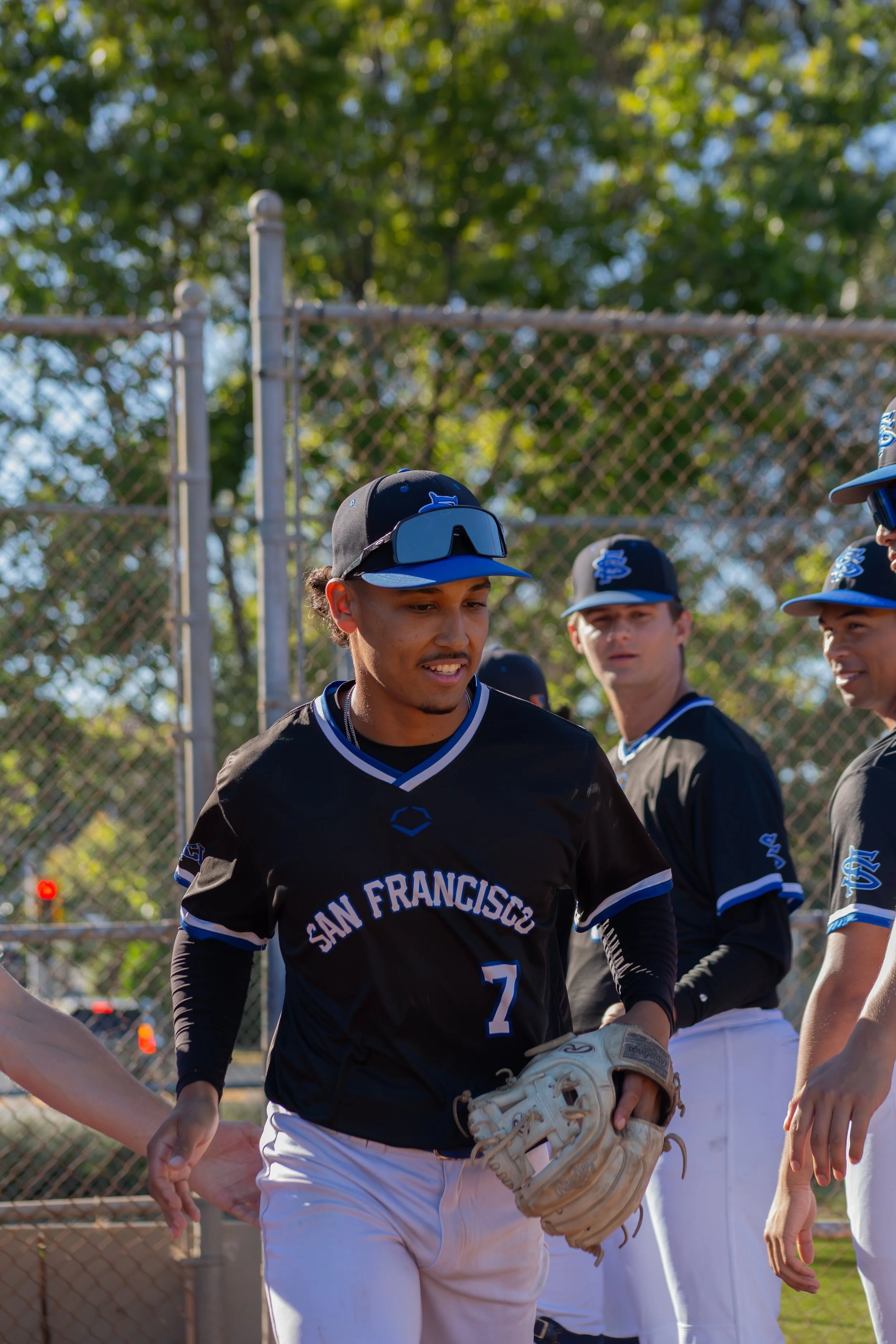 Young baseball players in black and white uniforms with blue accents, standing together on a sunny baseball field, one in the foreground wearing a glove