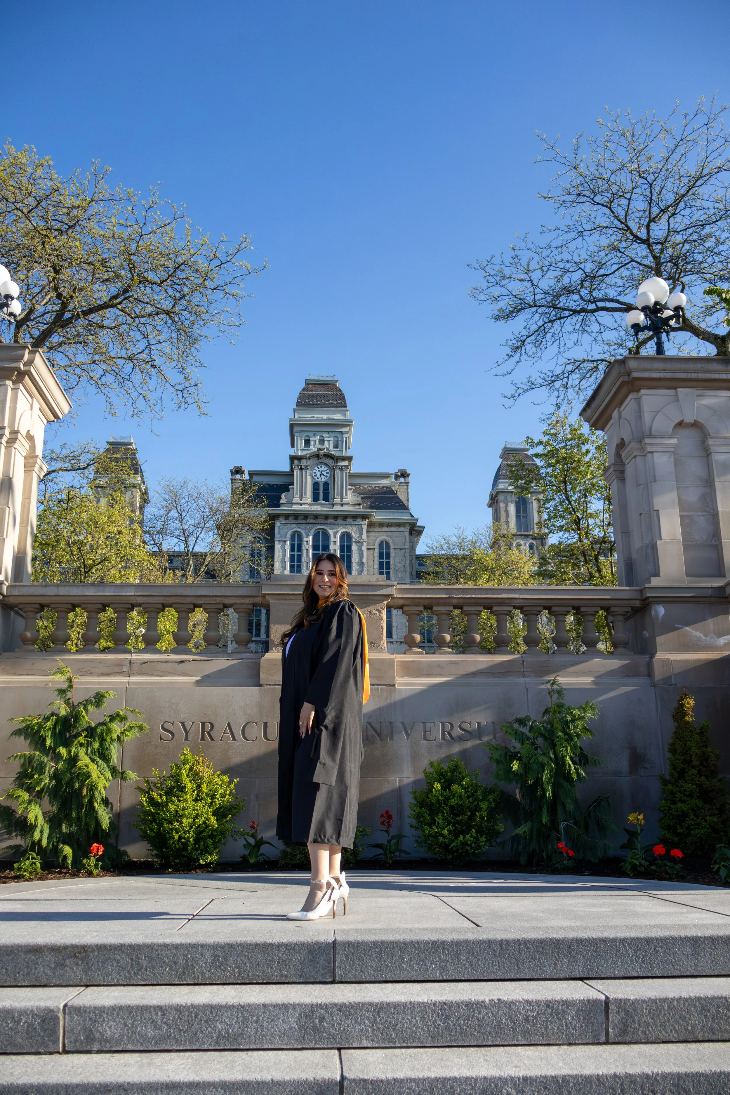 A woman in graduation attire standing in front of Syracuse University campus with trees, a historic building, and blue sky in the background.