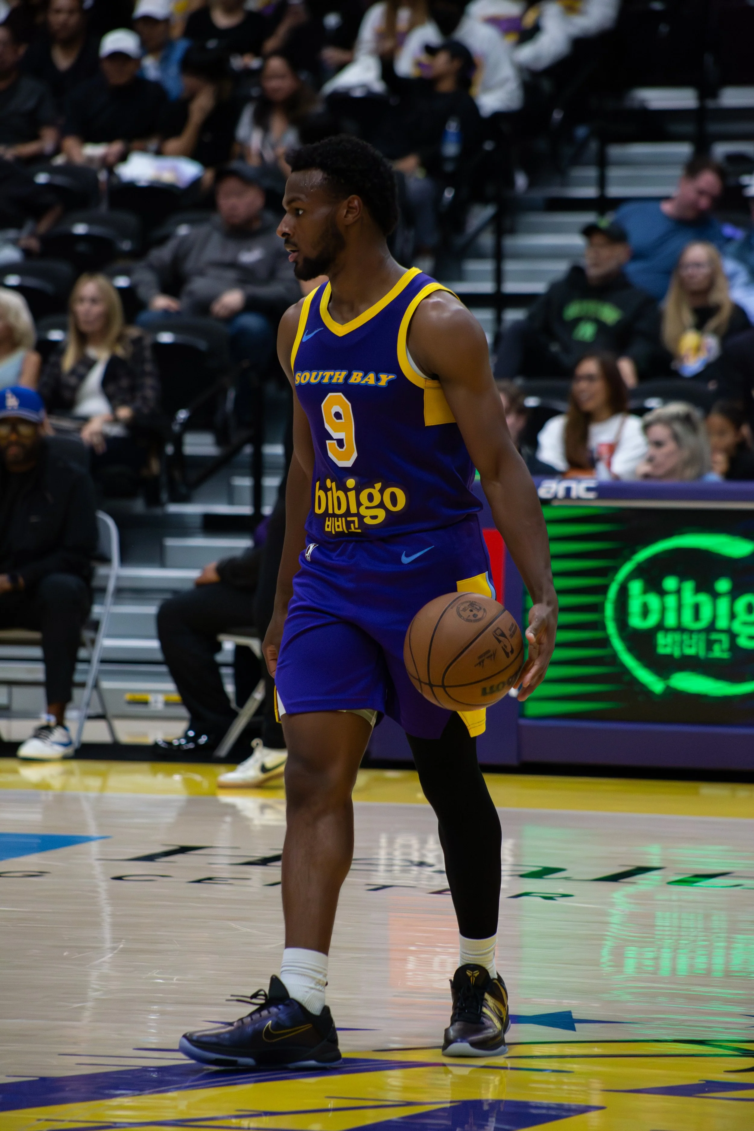 A basketball player in a blue and yellow uniform with the number 9 and the words 'South Bay' and 'bigo' on it, holding a basketball on the court during a game with spectators in the background.