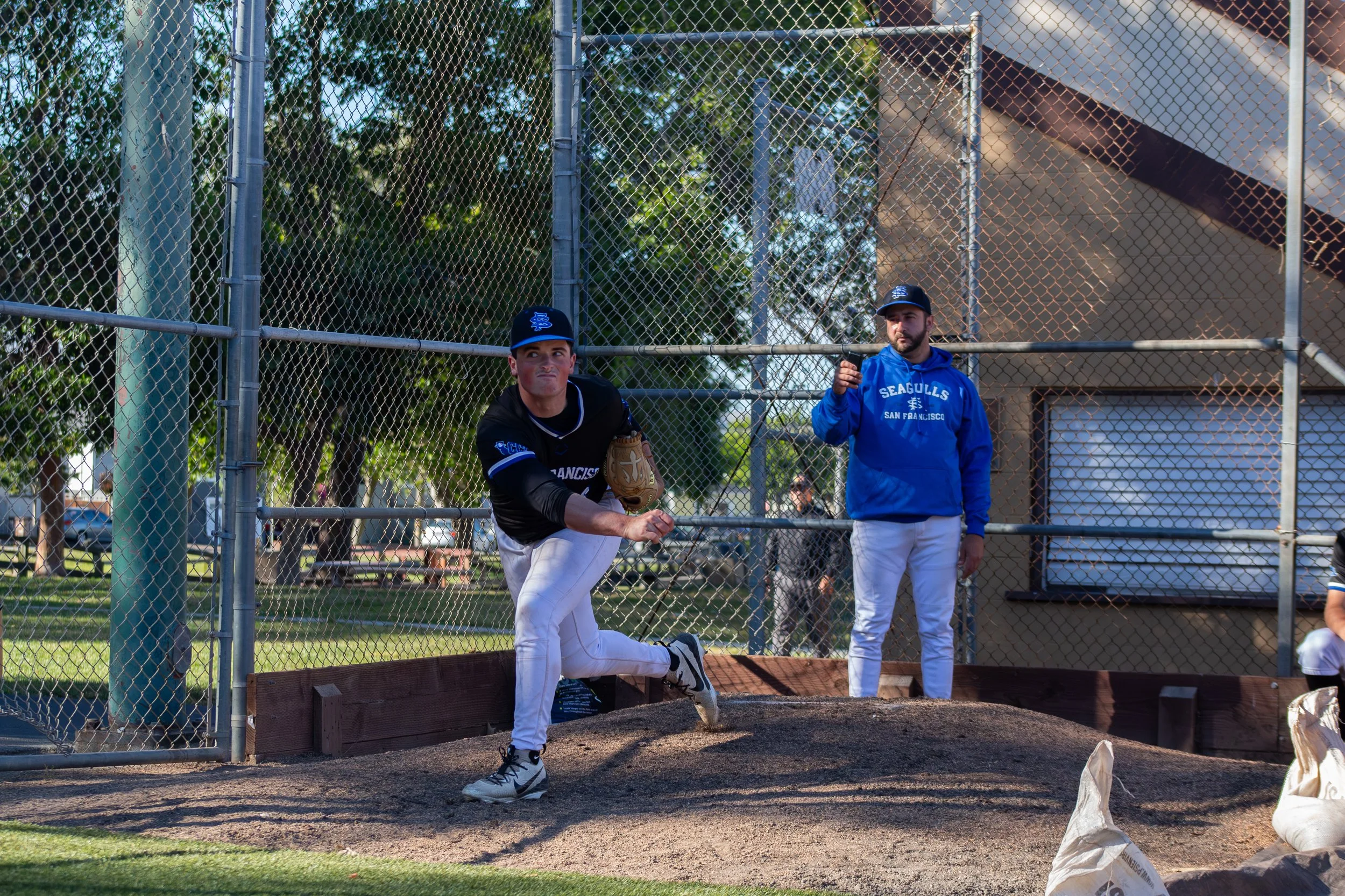 A baseball player in a black uniform with white pants and a blue cap is kneeling on a bench on a baseball field, preparing to pitch. Another person in a blue hoodie and white pants is standing nearby. The scene is viewed through a chain-link fence, w