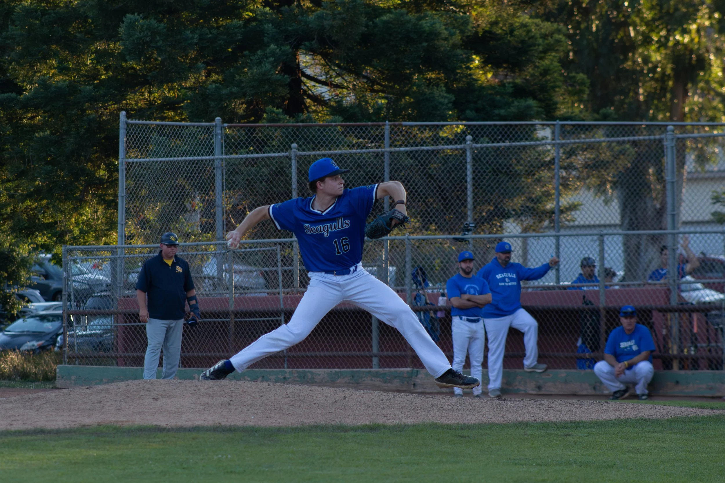 A baseball player in a blue uniform with the number 16 is pitching on a baseball field, with spectators and team members watching behind a chain-link fence, and trees in the background during late afternoon.