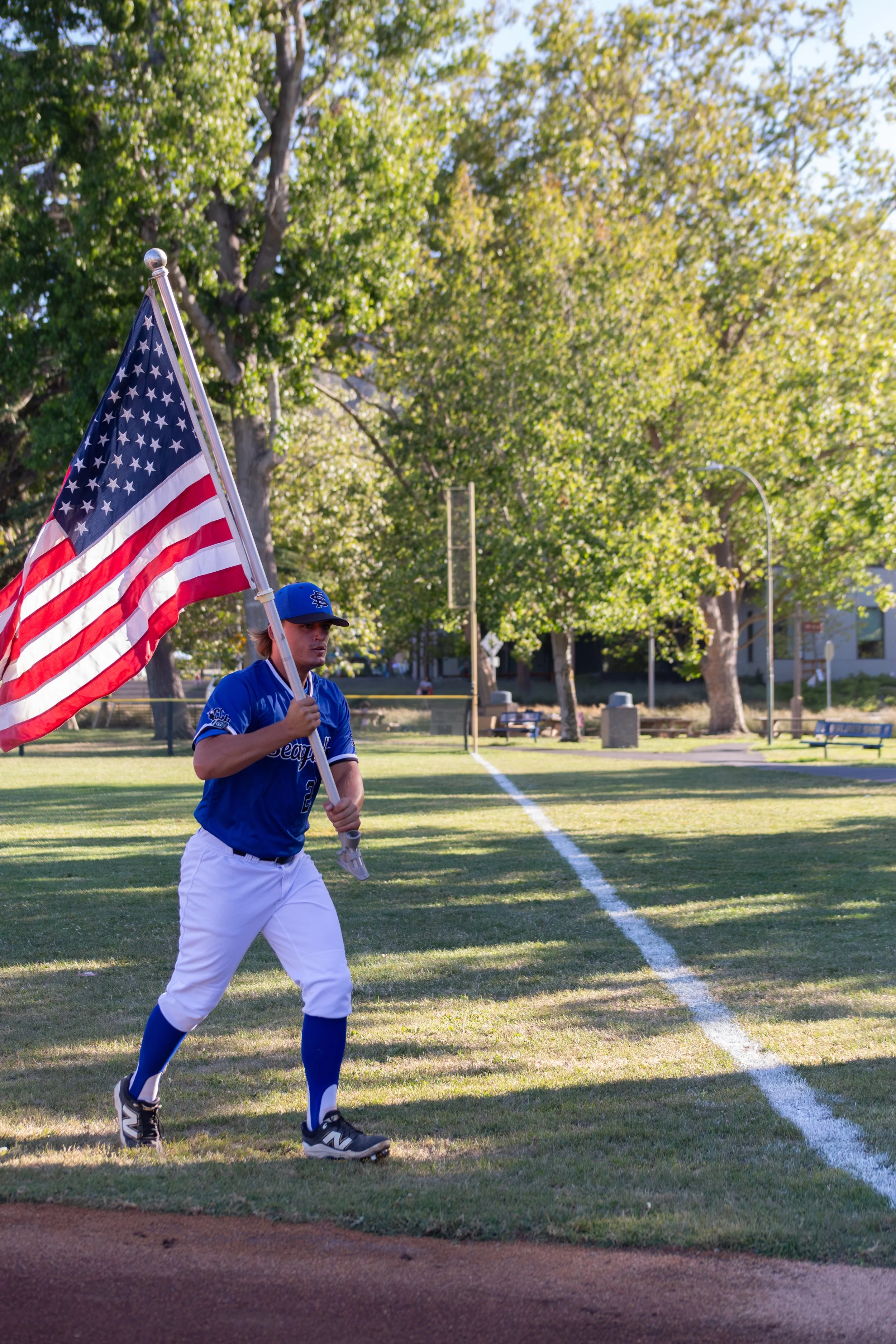 A baseball player in a blue uniform holding an American flag, walking on the field, with trees and benches in the background.