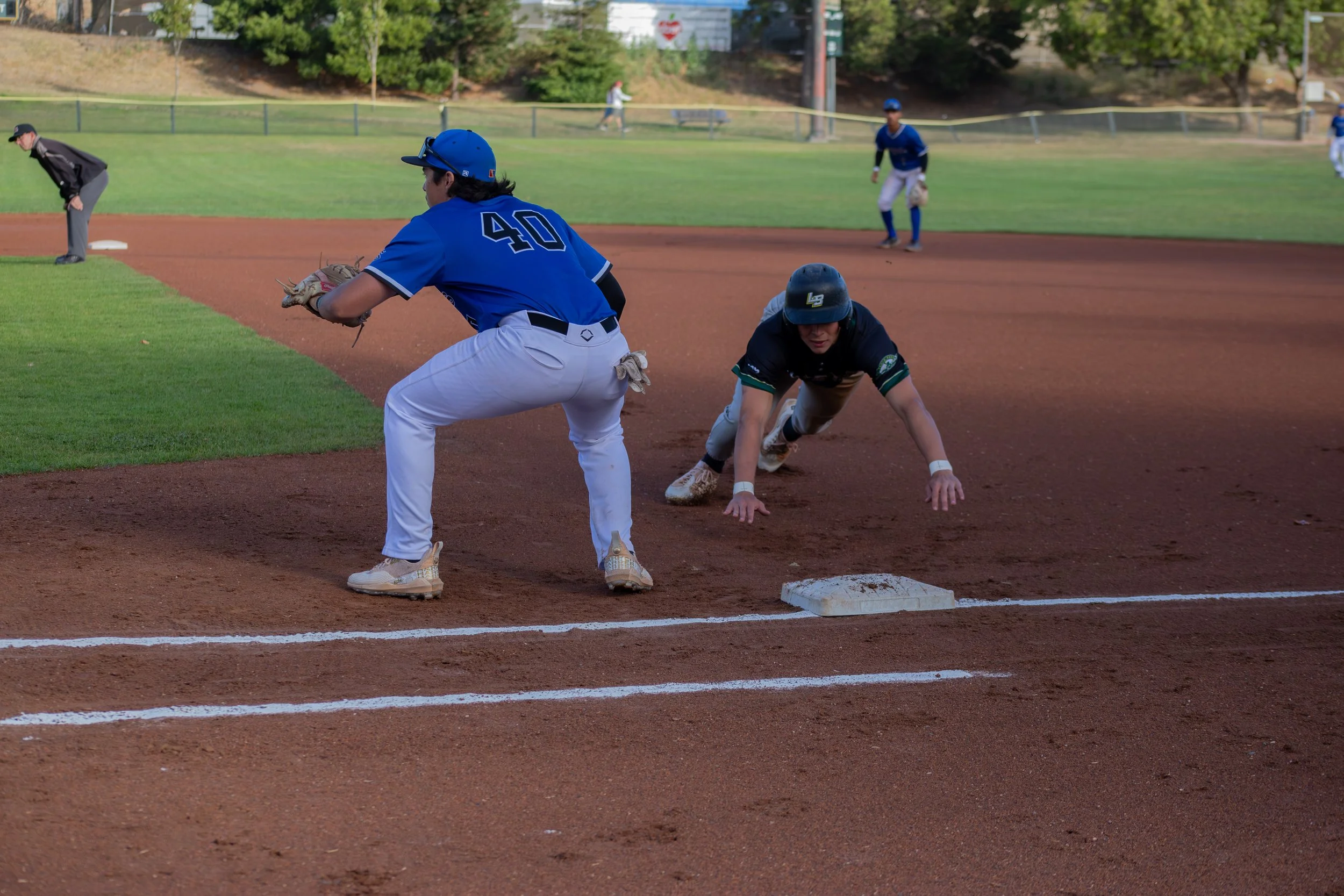 A baseball game in progress with players on the field, including a player in a blue jersey and white pants near the base, a fielder in a black helmet and dark uniform, and others in the background. The scene is set on a baseball diamond with grass an