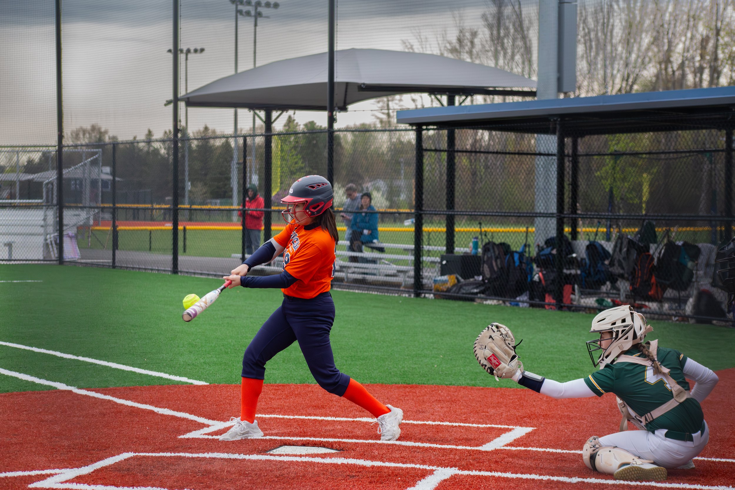 A young girl wearing a helmet and orange sports uniform swinging a yellow softball with a bat on a softball field, with another girl in catcher's gear kneeling behind her.