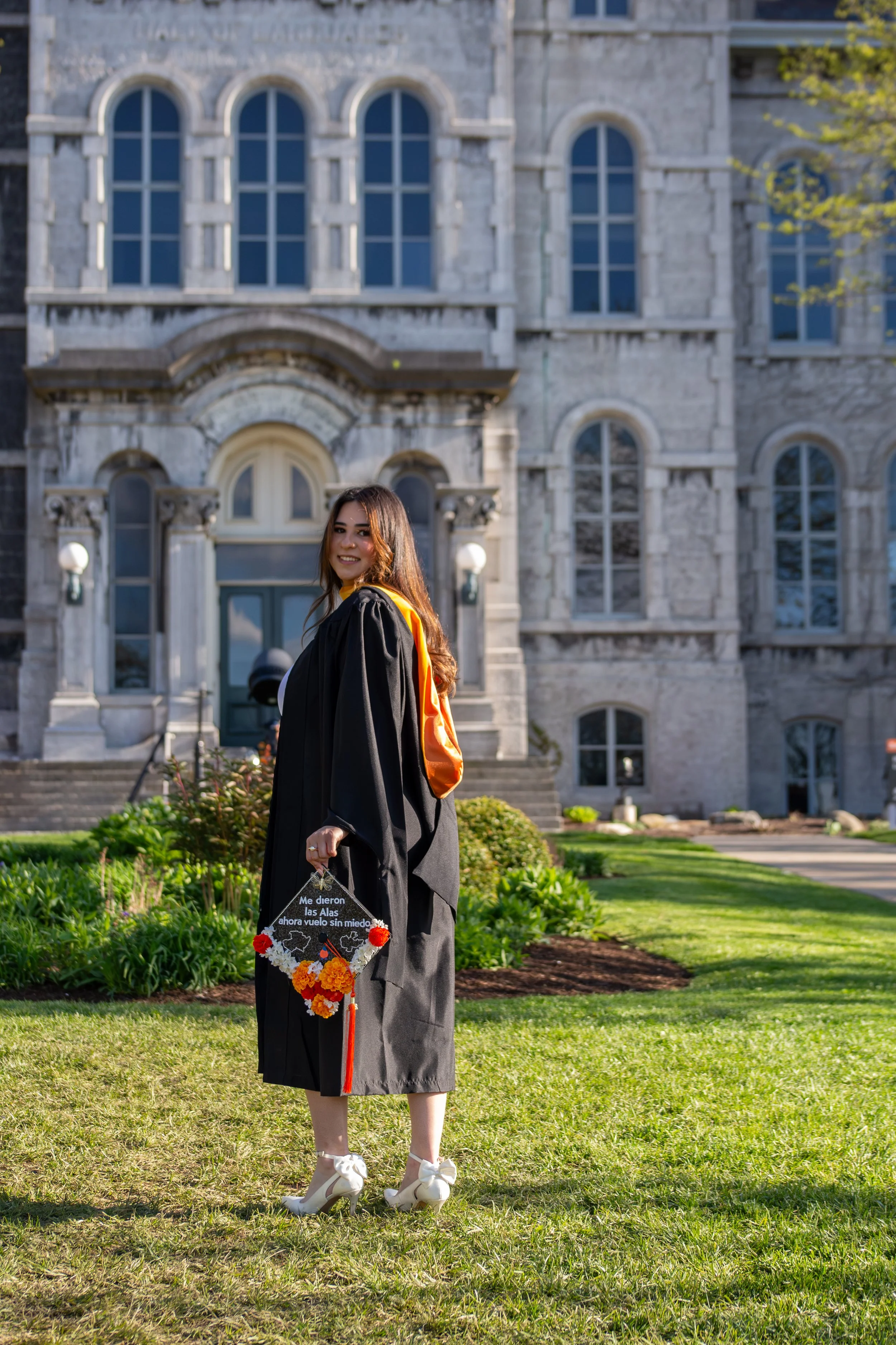 A young woman in a graduation cap and gown standing on a lawn in front of a large stone building with multiple arched windows. She is holding a decorated graduation cap and smiling.