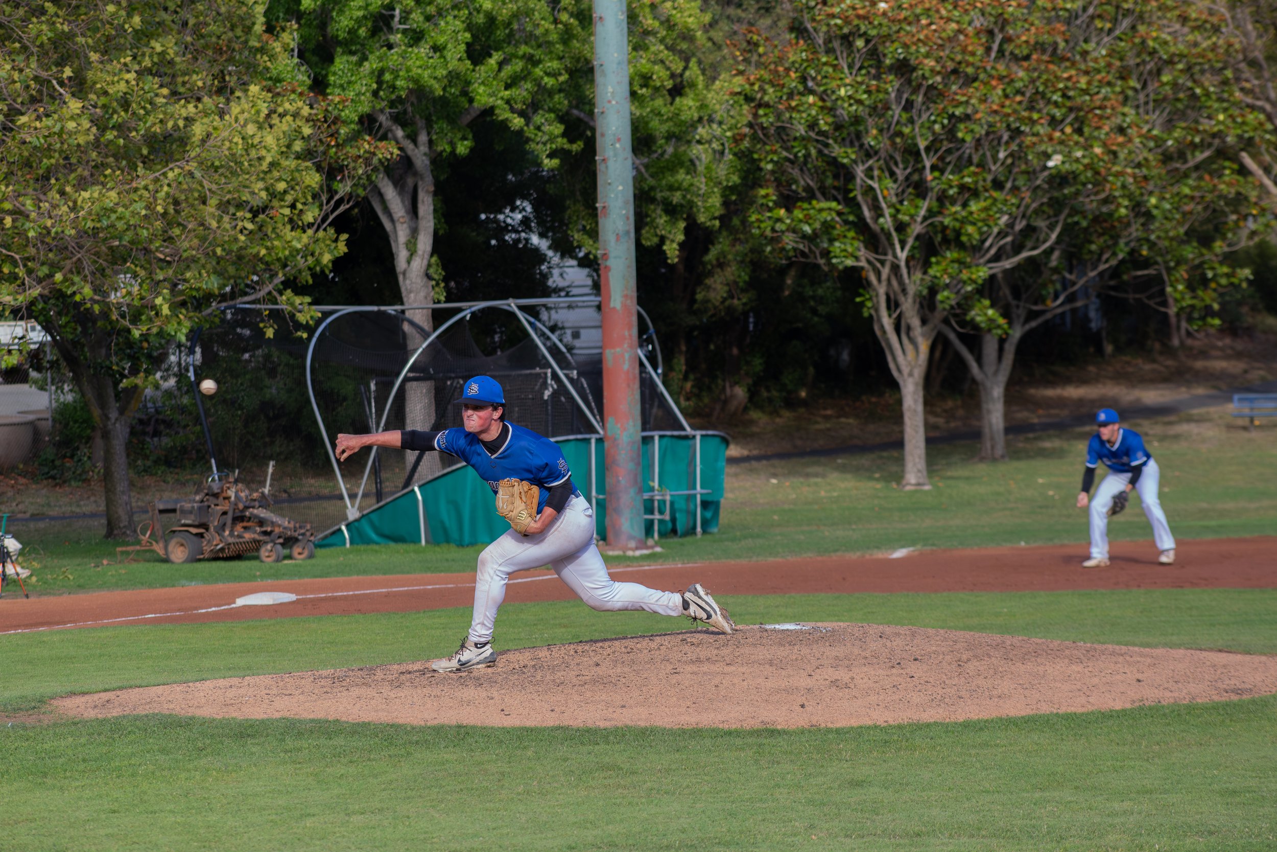 A baseball player in a blue uniform and cap is pitching a ball on a baseball field, with a teammate in the background preparing to catch or field the ball, surrounded by green trees.