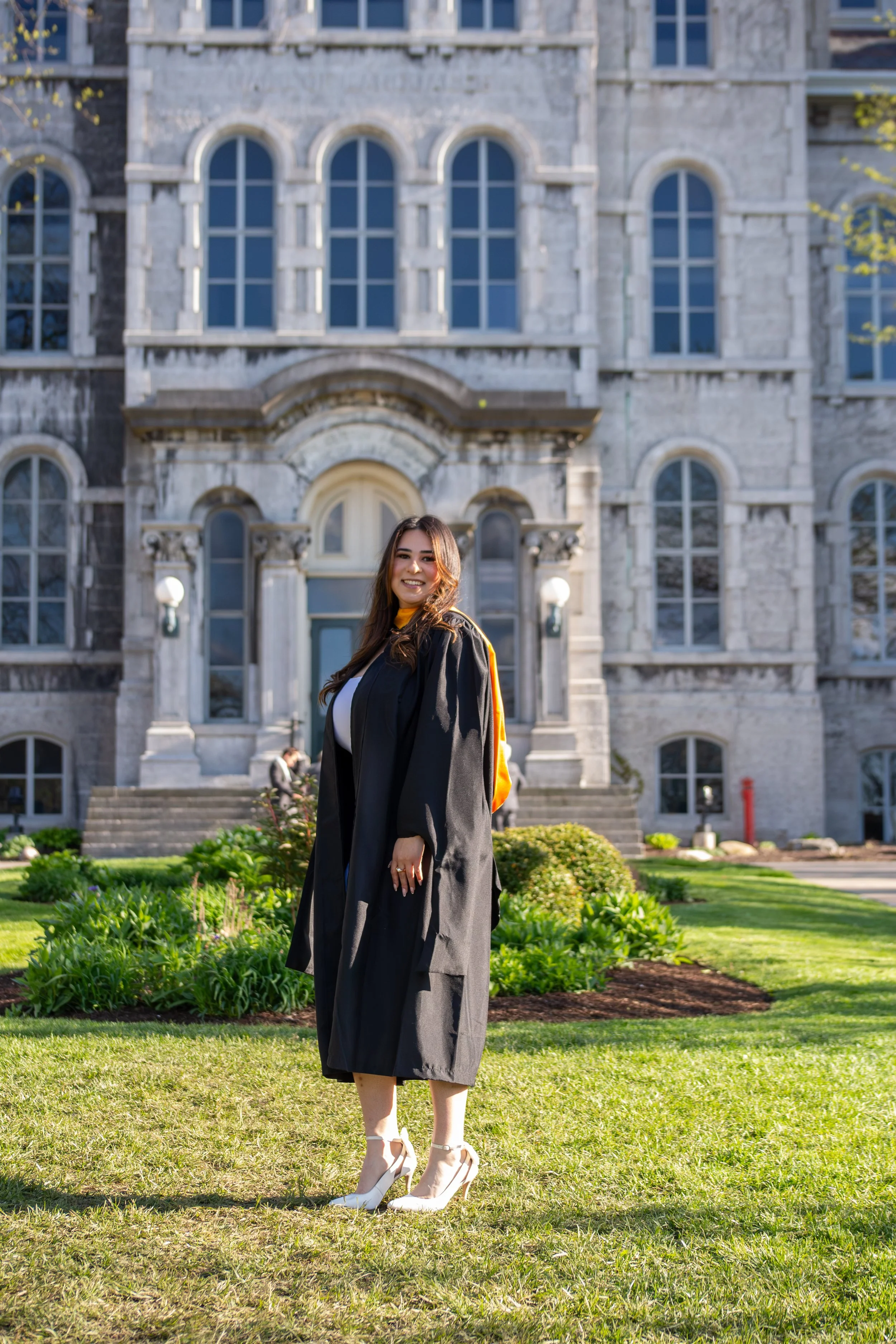 Young woman in graduation gown smiling outdoors in front of a historic stone building with large arched windows, well-kept garden, and sunlight.