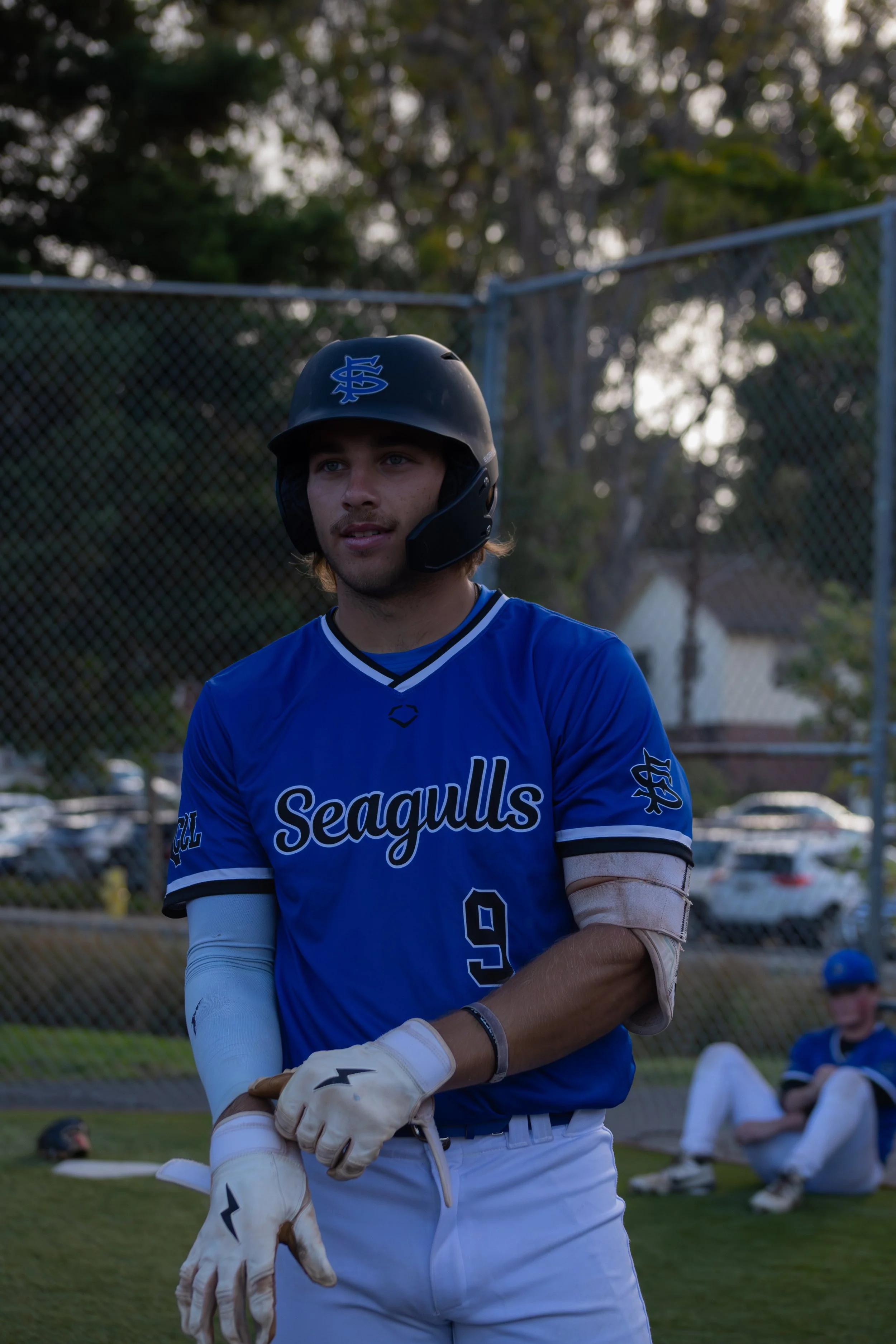 A baseball player wearing a navy helmet and a blue jersey with the word 'Seagulls' stands on a field, with trees and other players in the background.