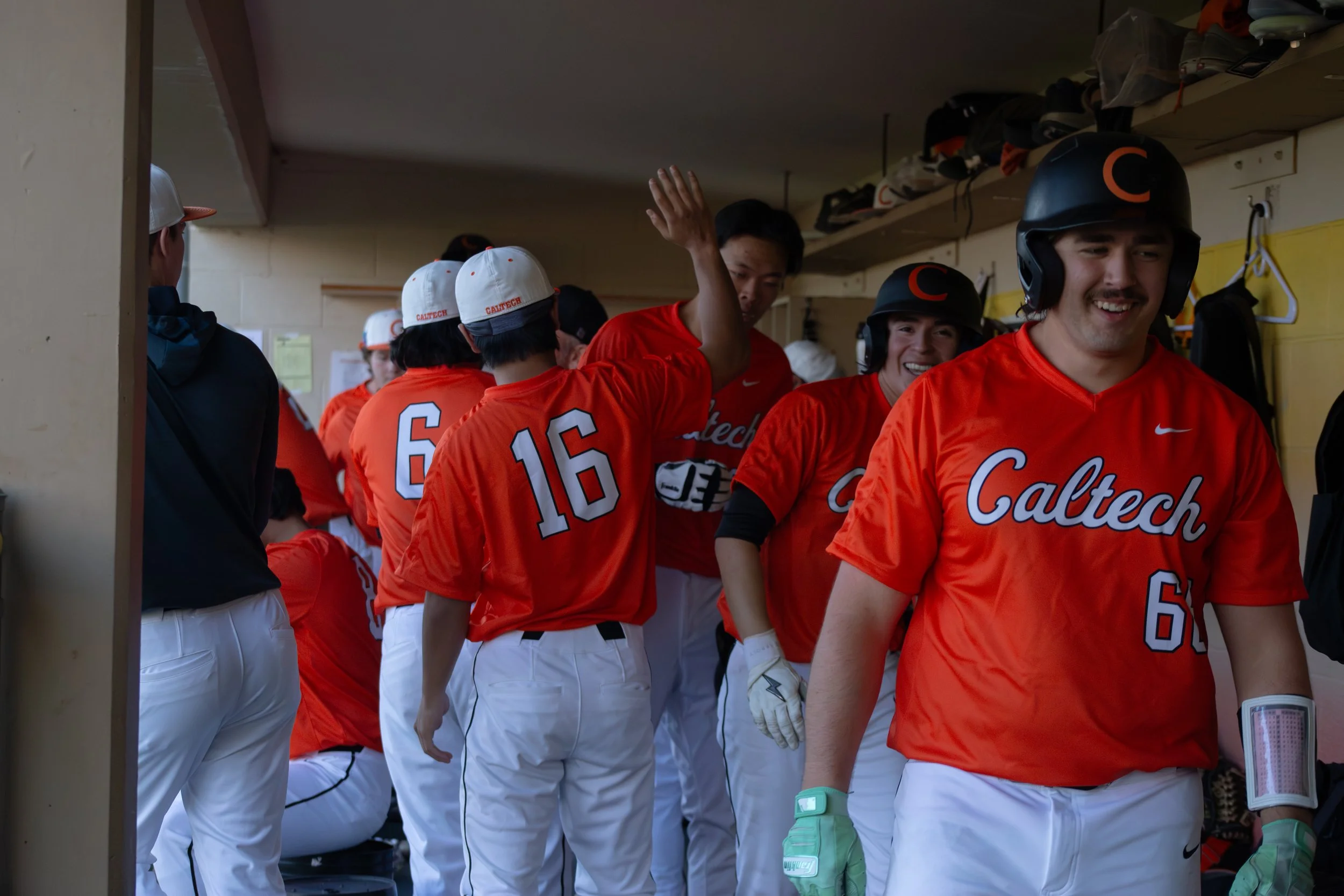 A group of baseball players in orange uniforms gather in a dugout, some wearing helmets and others with hats, preparing for a game or practice, and one player is waving.