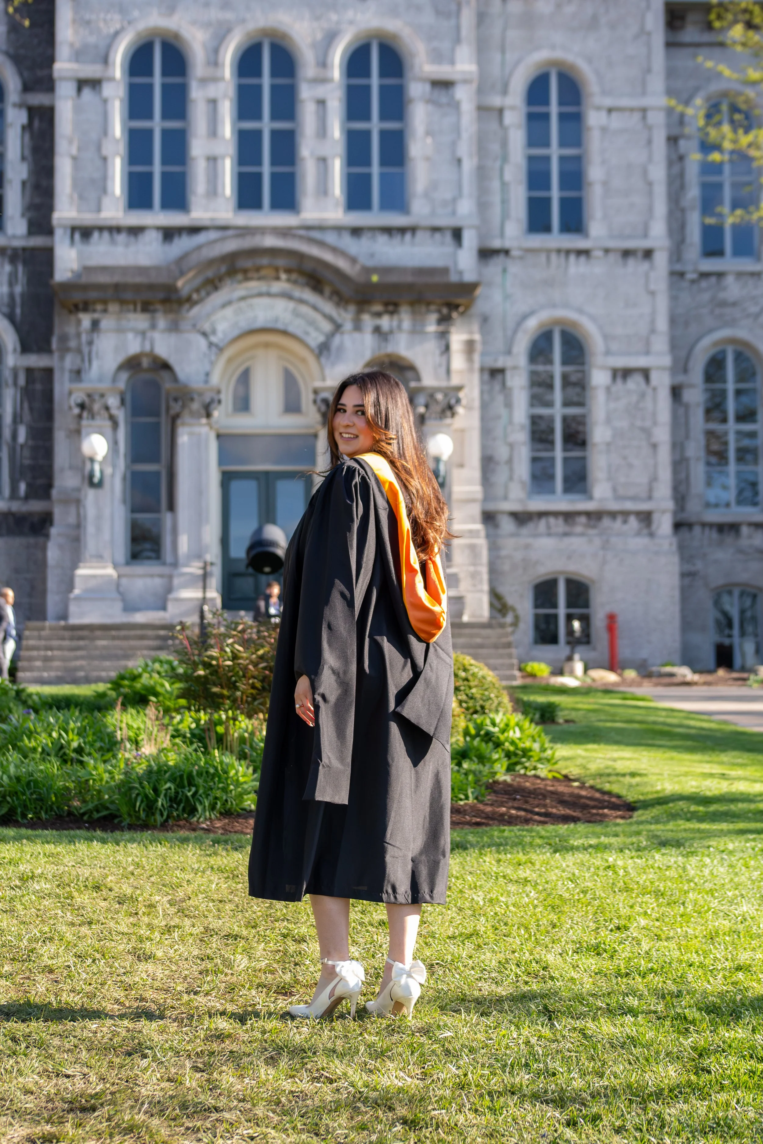A young woman in a graduation gown and cap stands outdoors on a green lawn, in front of a large, ornate stone building with arched windows, smiling.
