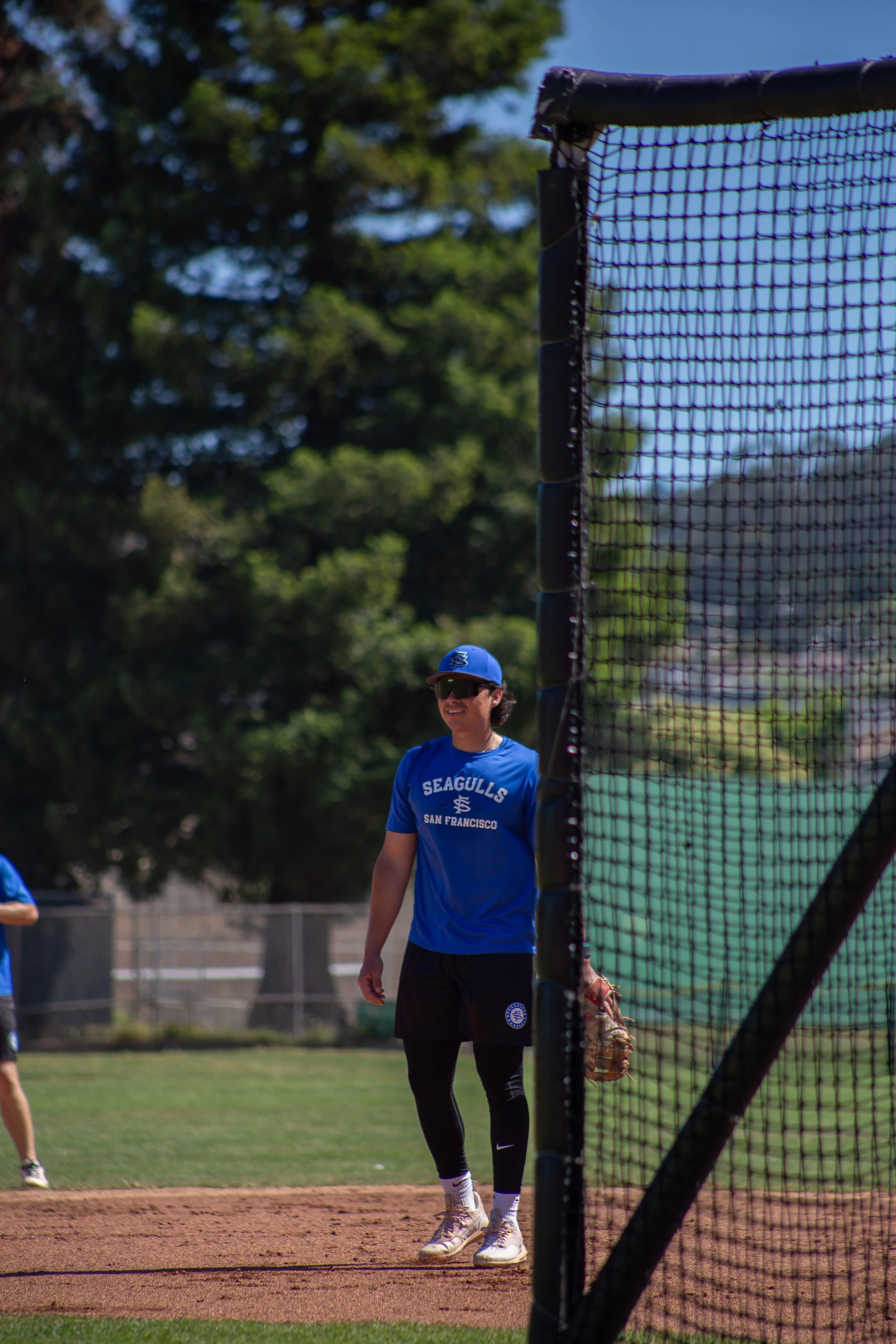 Person standing on a baseball field behind a fence, wearing sunglasses, a blue shirt with 'Seagulls San Francisco' logo, black shorts, and white sneakers, with a baseball glove in hand, near the baseball net.