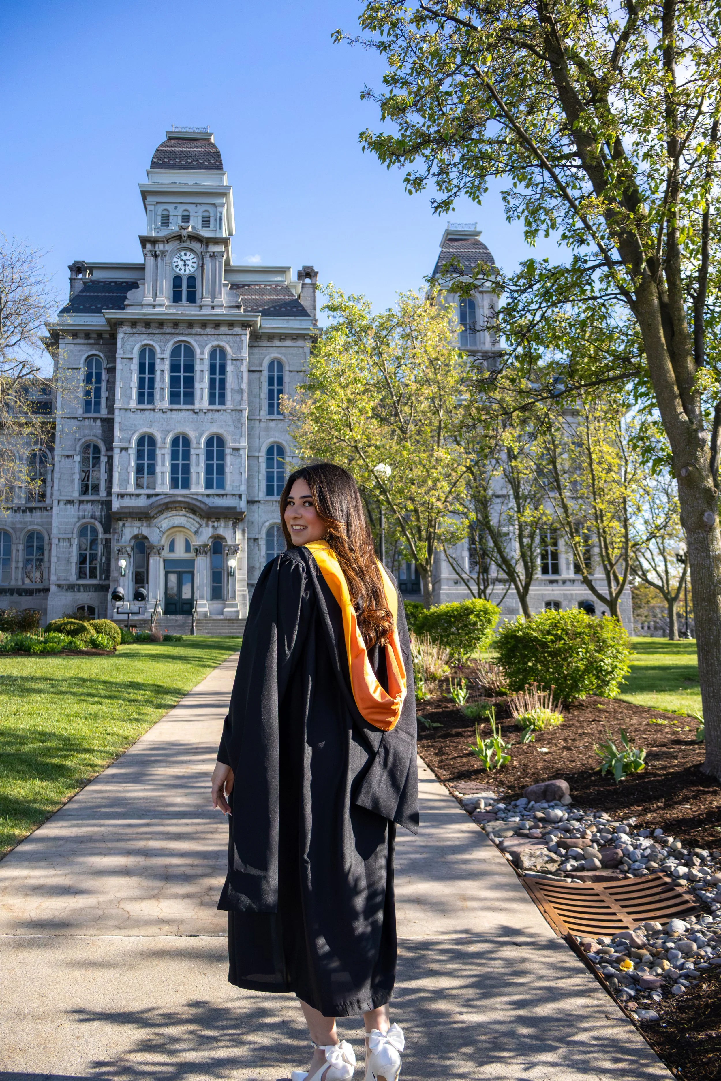 Young woman in graduation gown and cap walking on campus with historic building and trees in background.