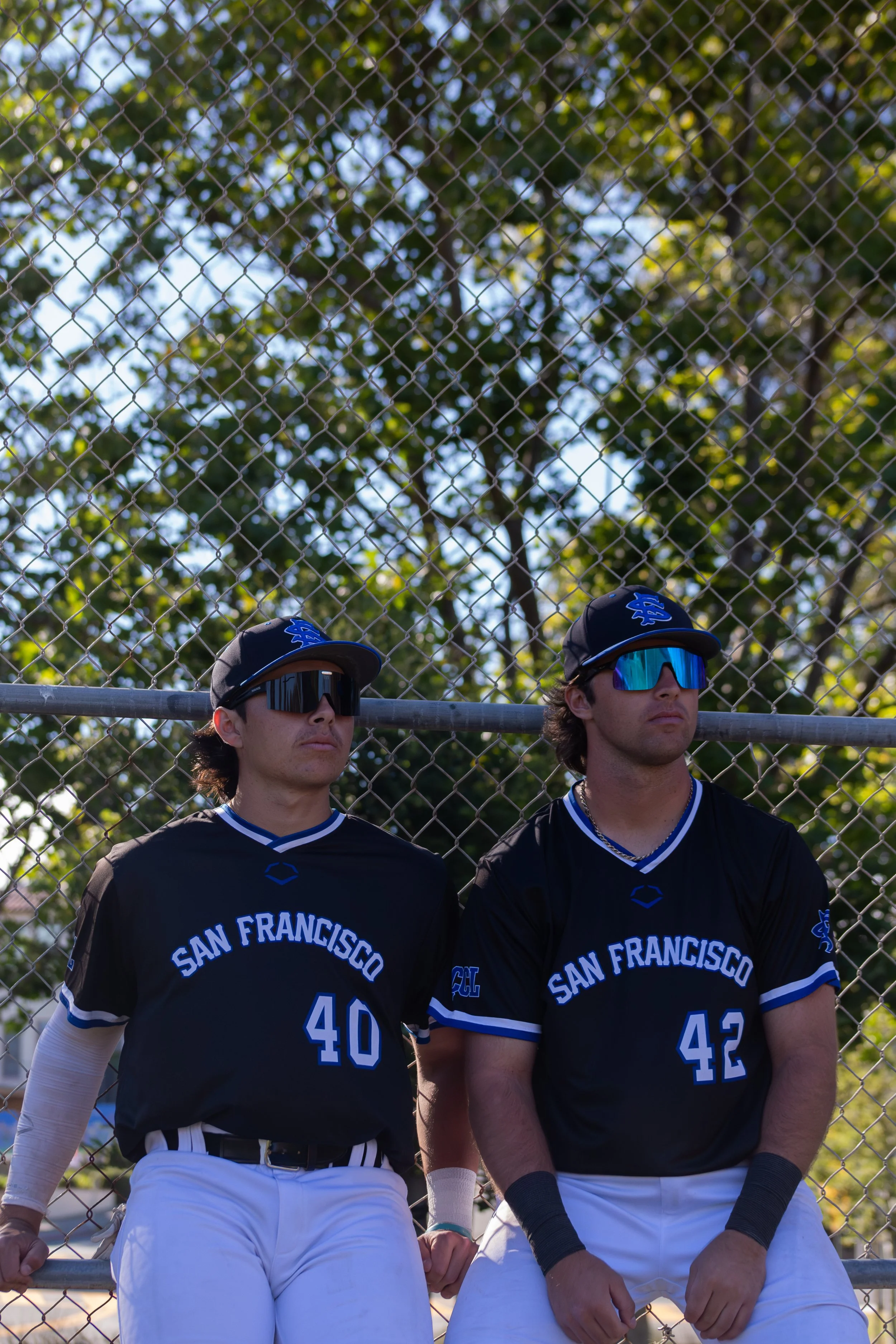 Two baseball players wearing black jerseys with blue and white accents and blue caps stand in front of a chain link fence with trees behind them. They are looking slightly upward, wearing sunglasses, and appear serious.