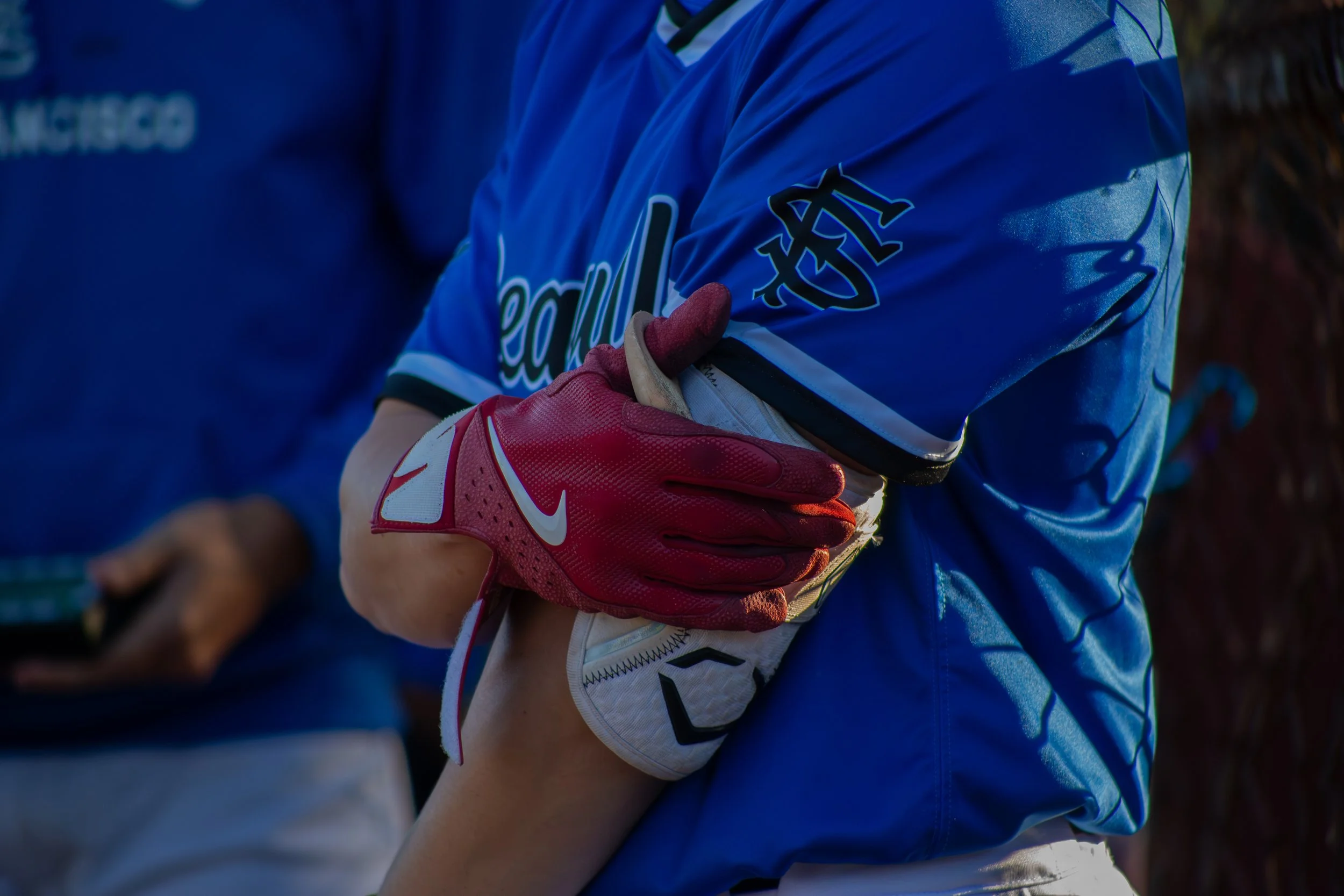 Close-up of a person wearing a blue sports jersey and red gloves, holding their gloved hand over their chest. The background is blurred, indicating an outdoor setting.