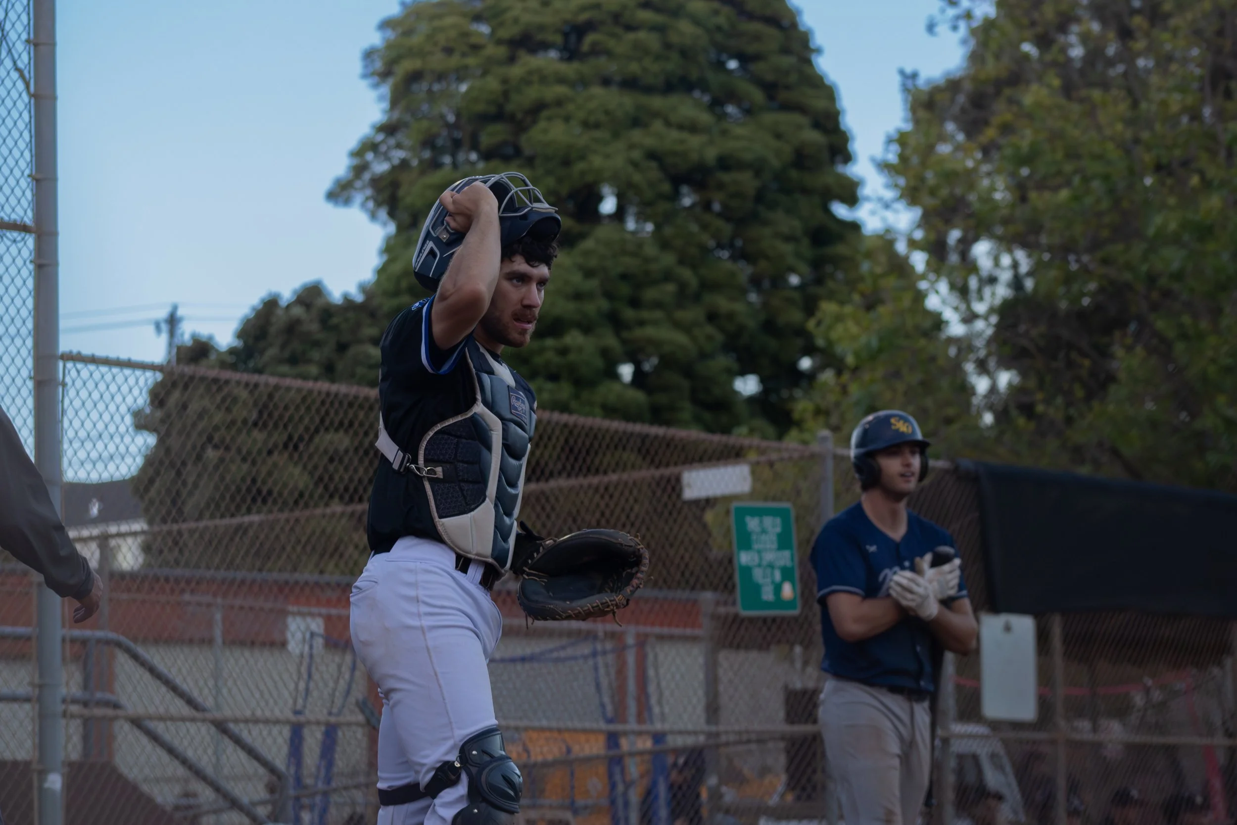 A young male baseball catcher adjusting his helmet on a baseball field with trees and a chain-link fence in the background.