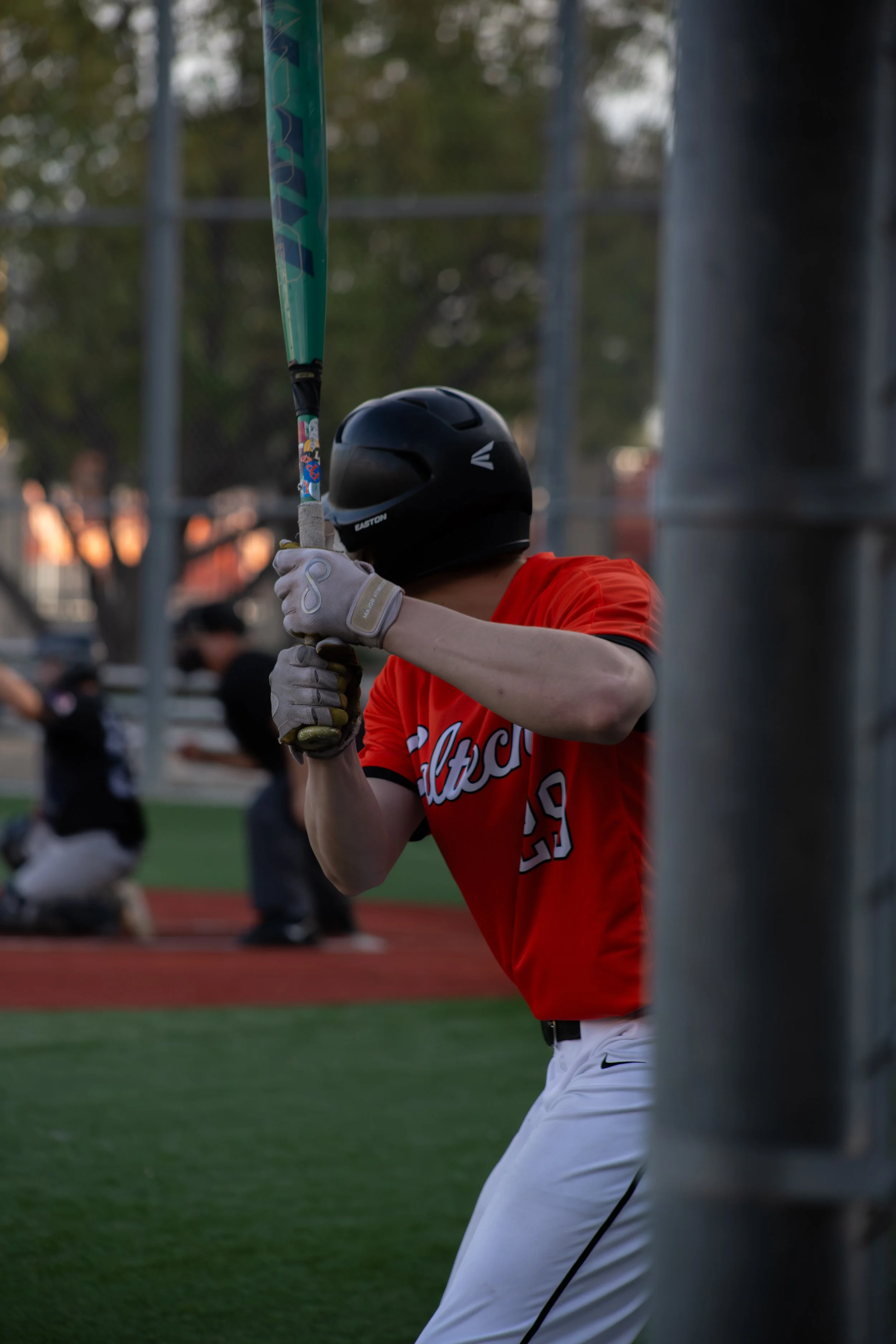 A baseball player in a red uniform and white pants, holding a bat, preparing to swing during a game at a baseball field, with a helmet and gloves, behind a chain-link fence.