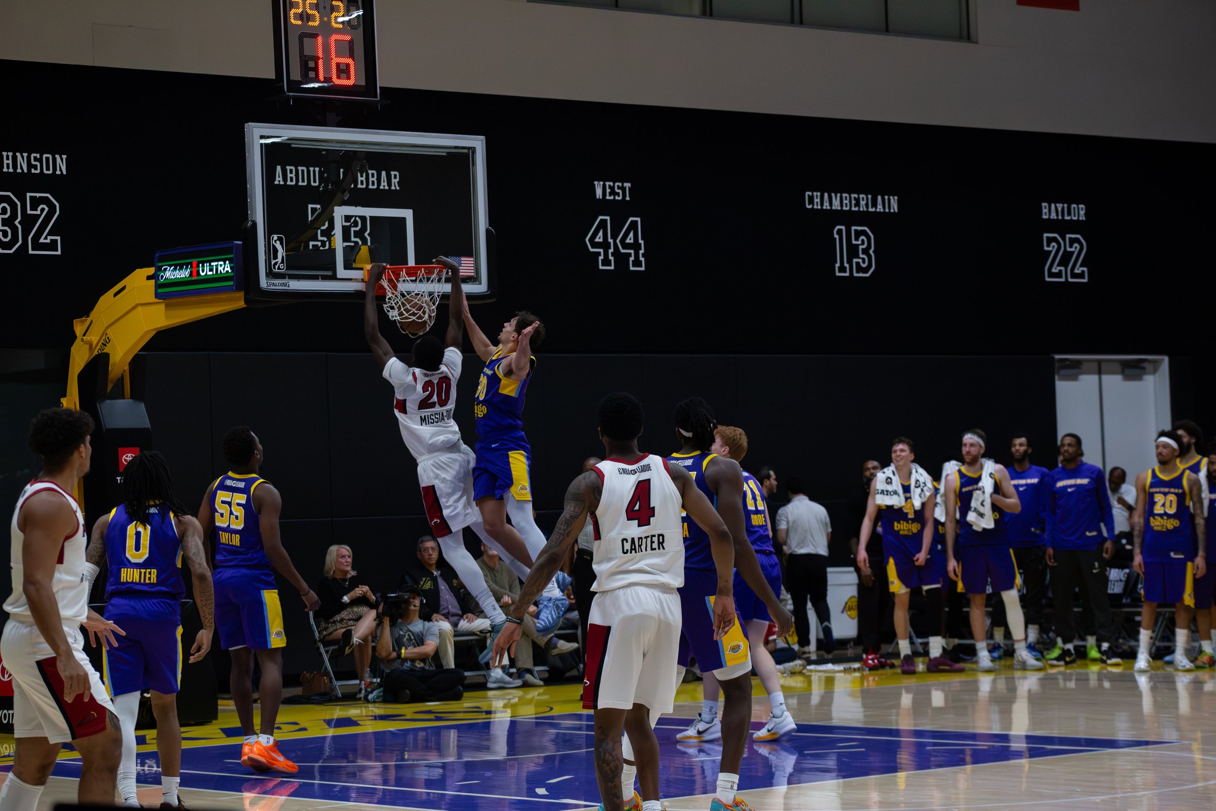 A basketball game in progress with players jumping for a rebound near the basket. The players are wearing red, white, blue, and yellow jerseys. Spectators are seated on the sidelines and watching the game.
