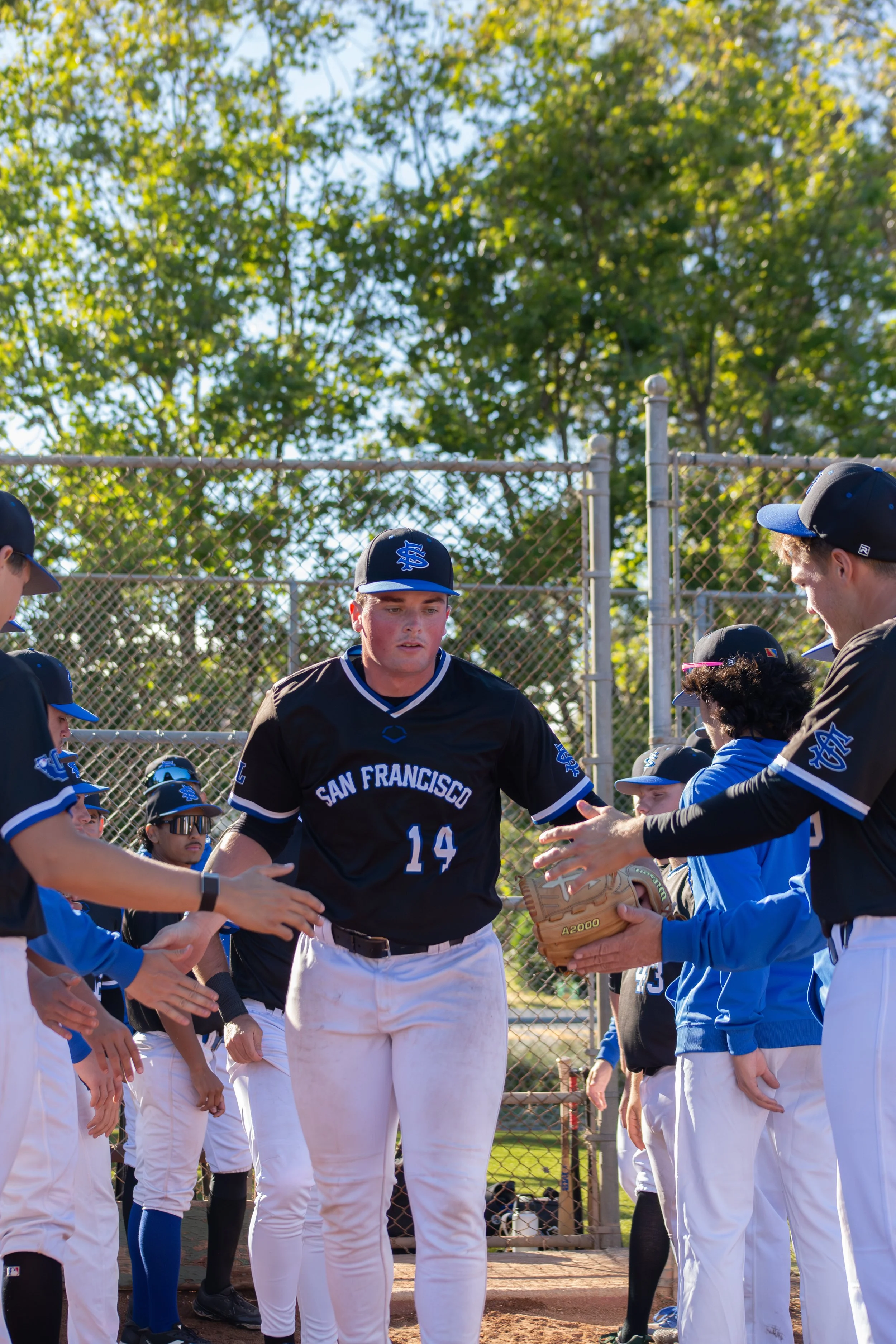 Baseball players in team uniforms standing in a line on a baseball field, with a coach or player in the center handing out a baseball glove, outdoors with trees in the background.