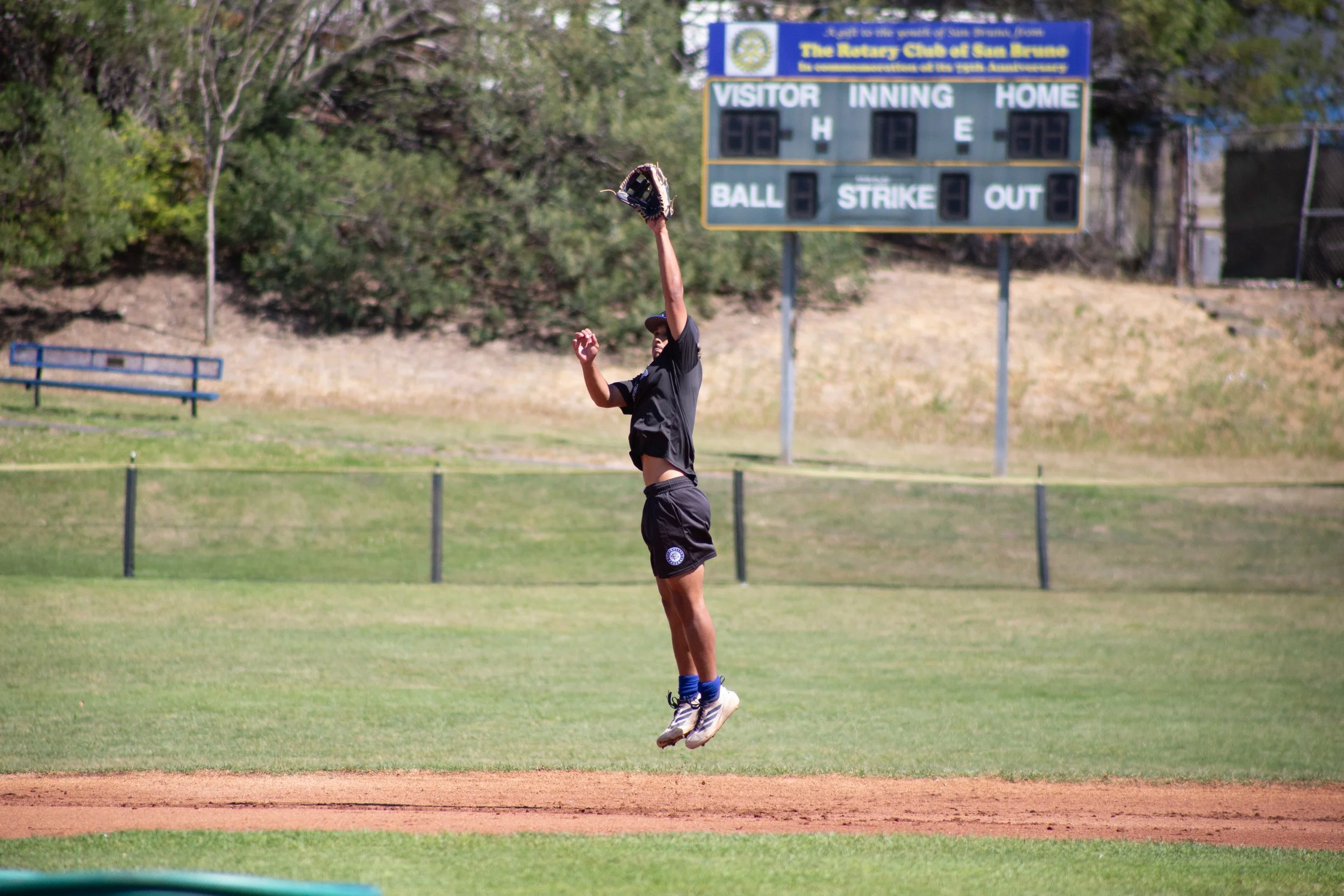 A person jumping to catch a ball on a baseball or softball field with a scoreboard in the background.