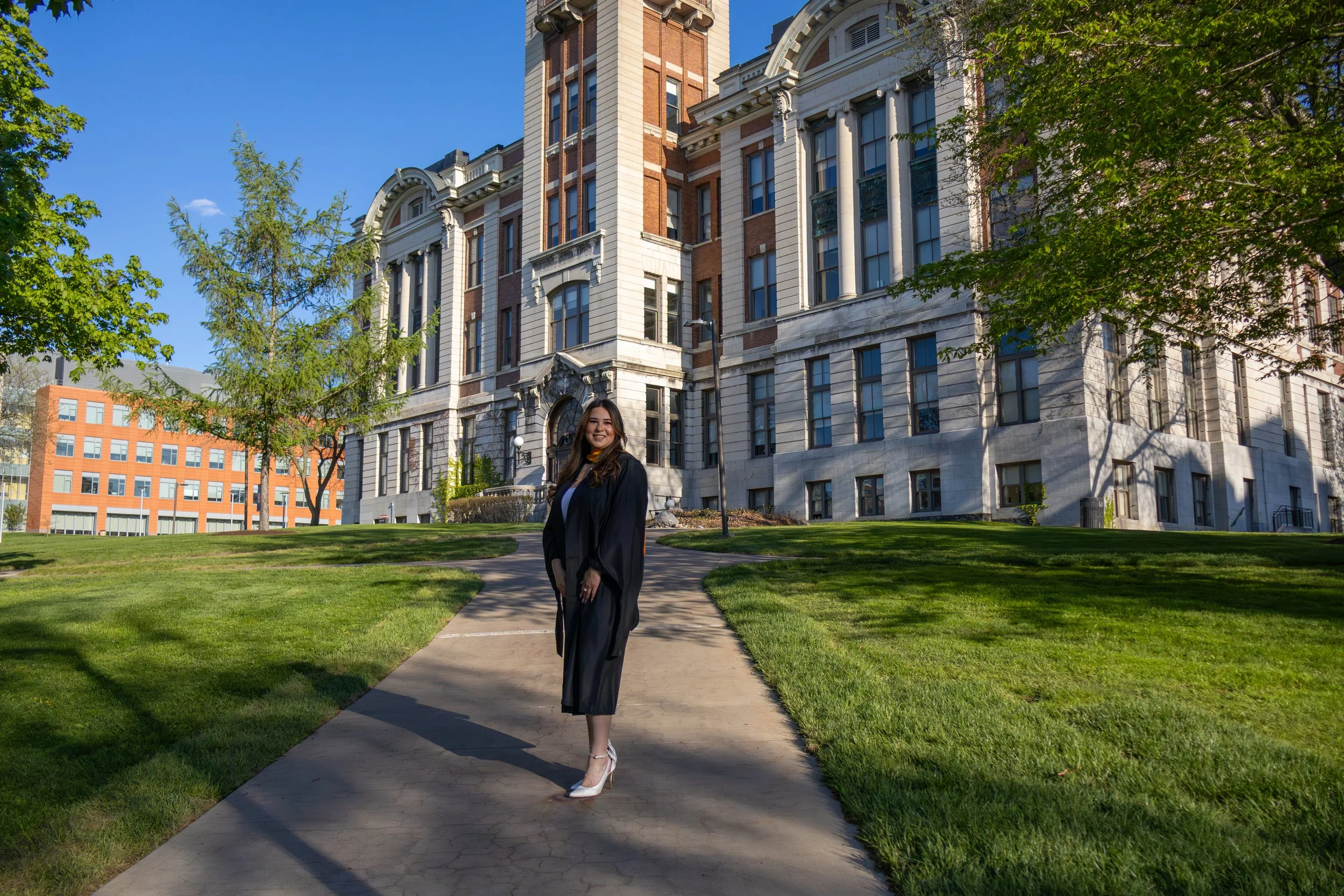 A young woman in a graduation cap and gown standing on a sidewalk in front of a historic building with large windows and columns, with green grass and trees around on a bright sunny day.
