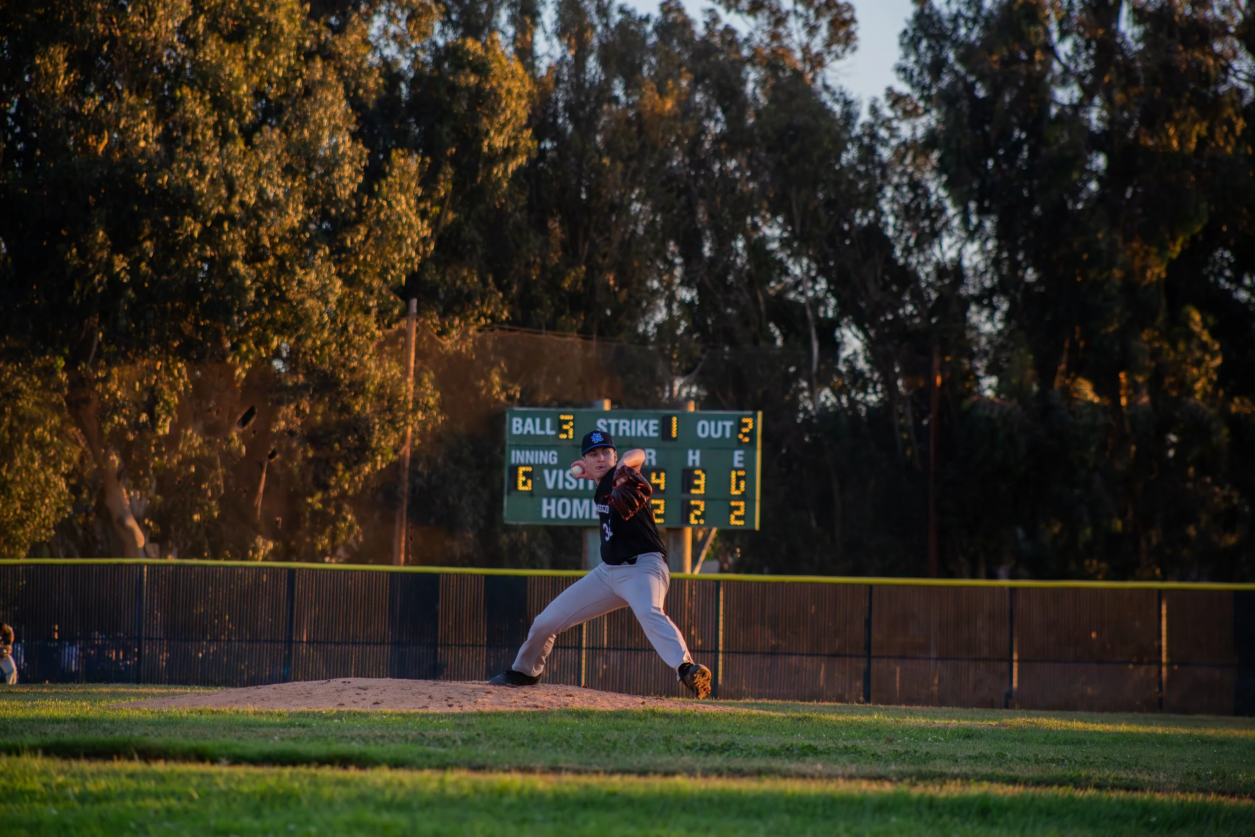 A baseball pitcher in a black and orange uniform is throwing a pitch on the mound at a baseball field during sunset, with trees and a scoreboard in the background.