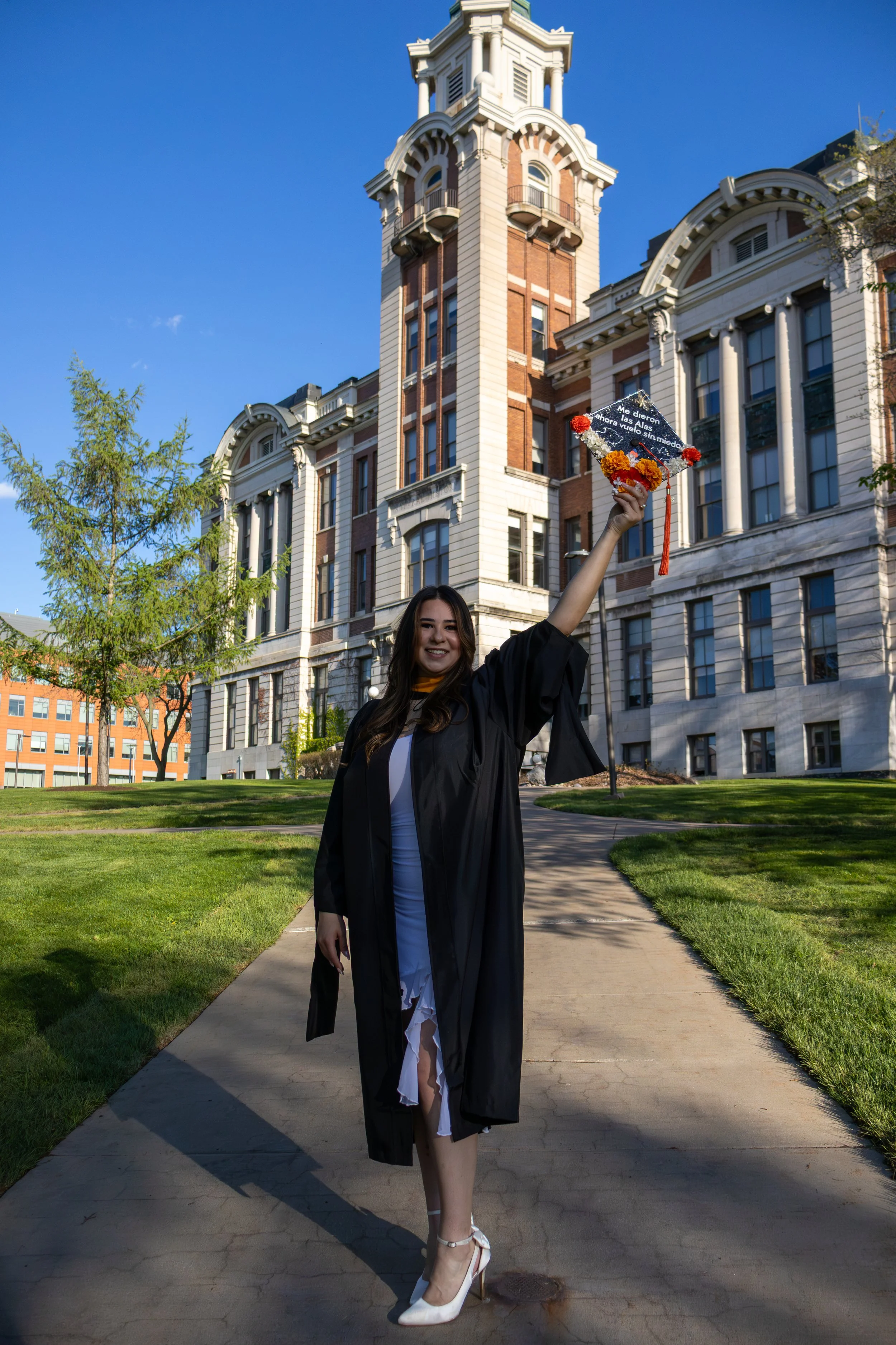 A young woman in a graduation gown and cap holding a decorated mortarboard in front of a historic building with a clock tower. She is smiling, standing on a sidewalk with green grass and trees around.