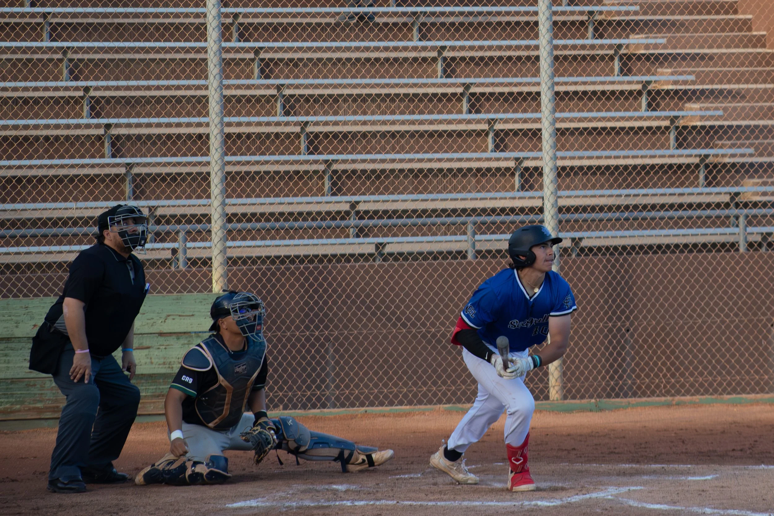 A baseball game scene with a female batter in a blue uniform awaiting a pitch, a catcher and umpire crouching behind her, and a chain-link fence in the background.