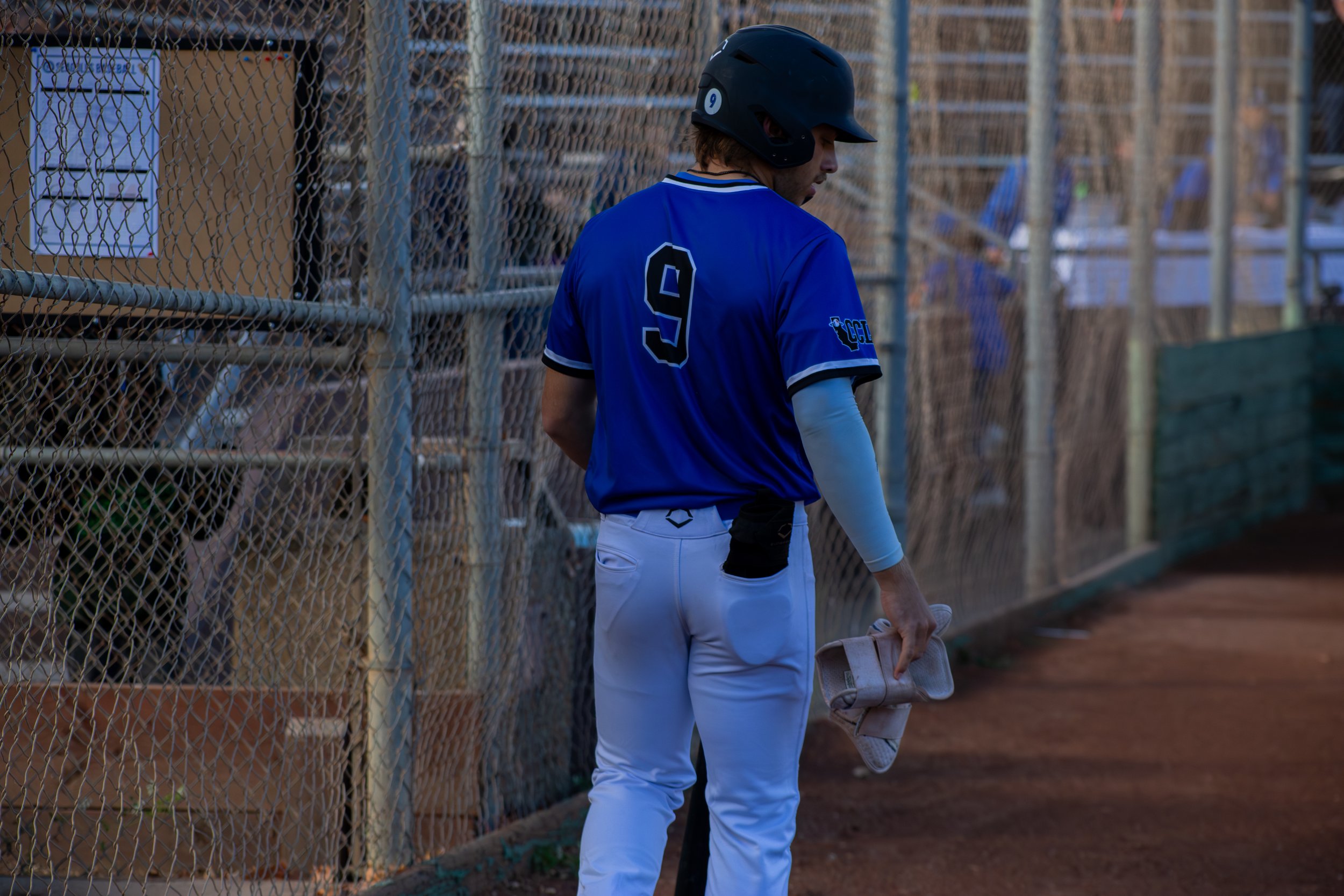 Baseball player dressed in a blue jersey and gray pants holding a baseball bat, standing near a chain-link fence at a baseball field.