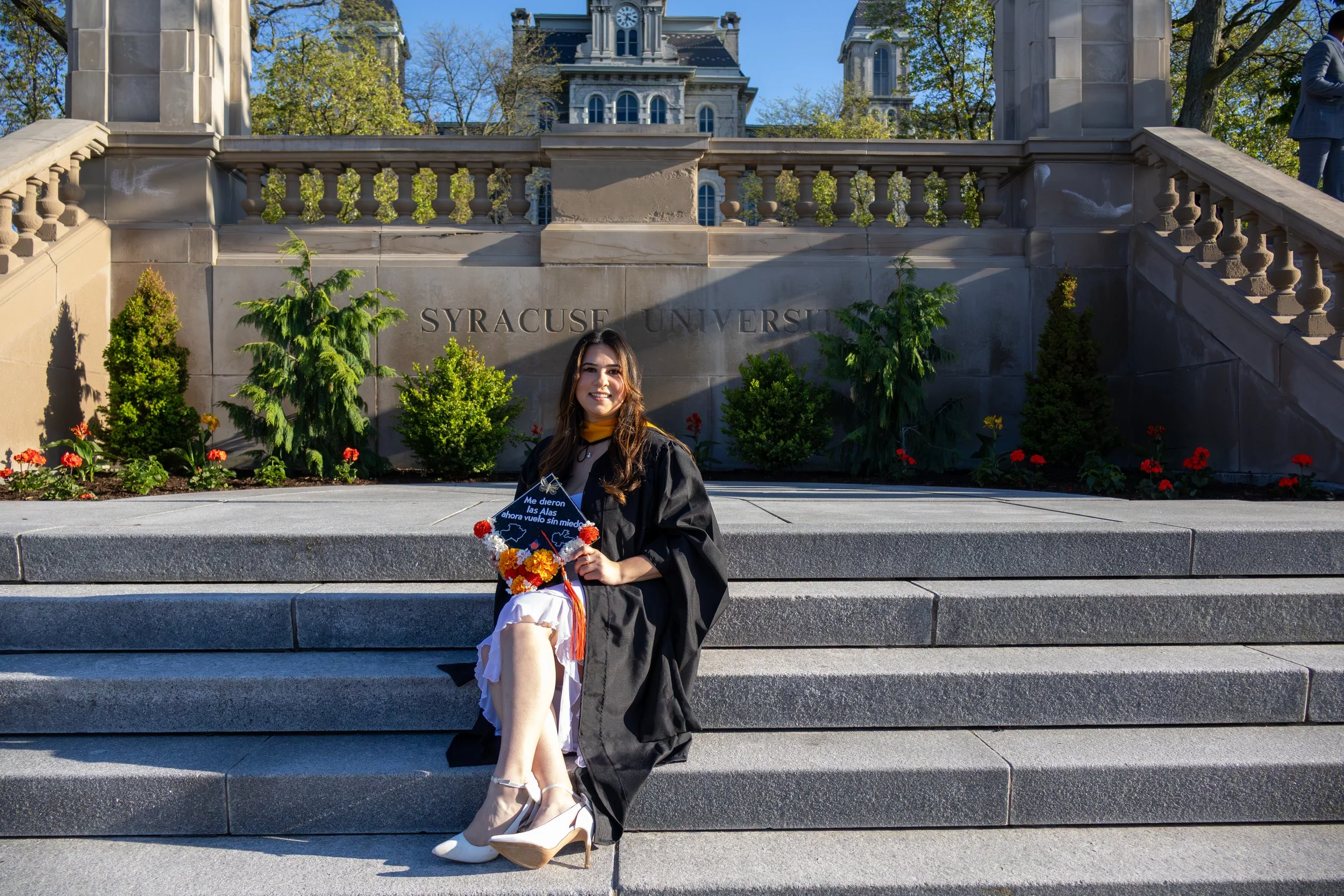 A young woman in a graduation gown and cap sitting on the steps of Syracuse University during graduation day, holding a certificate and a bouquet of flowers, with the university's building and trees in the background on a sunny day.