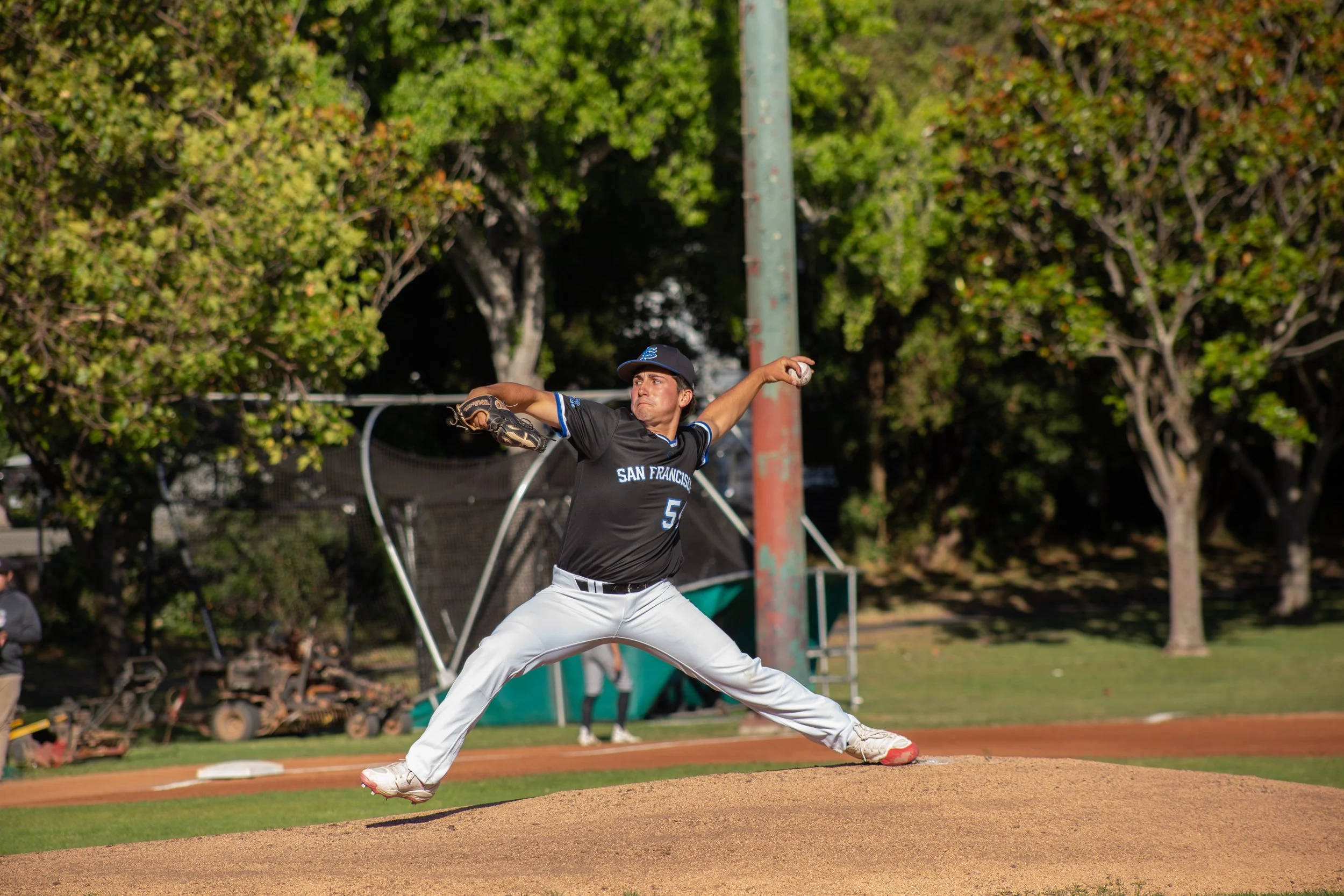 A male baseball pitcher in a black uniform with 'SAN FRANCISCO' written on it, mid-throw on a baseball field, with trees in the background.