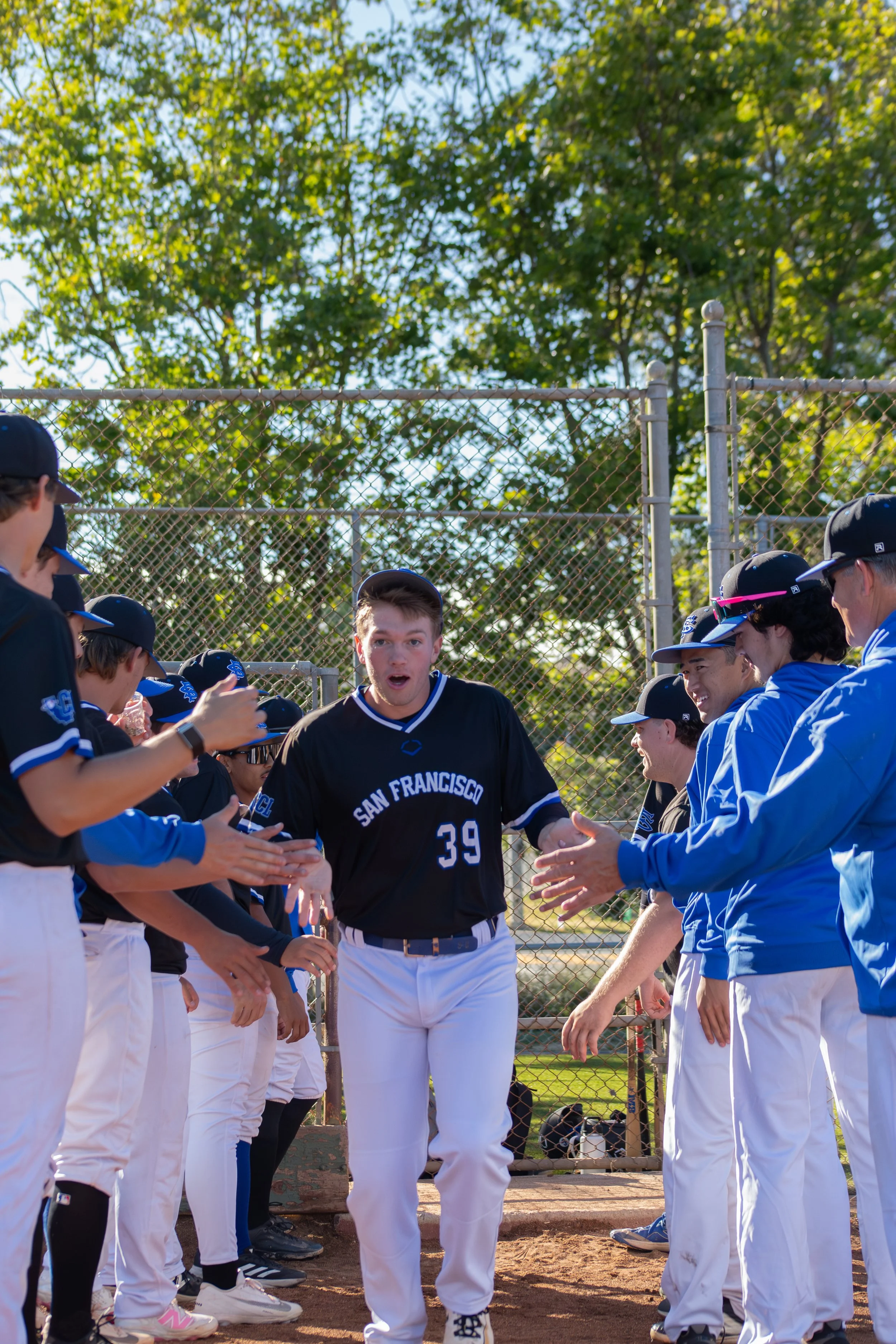 A baseball player in a black uniform with "San Francisco" written on it, standing between two lines of people who appear to be team members or fans, giving high fives in a baseball field with a chain-link fence and green trees in the background.