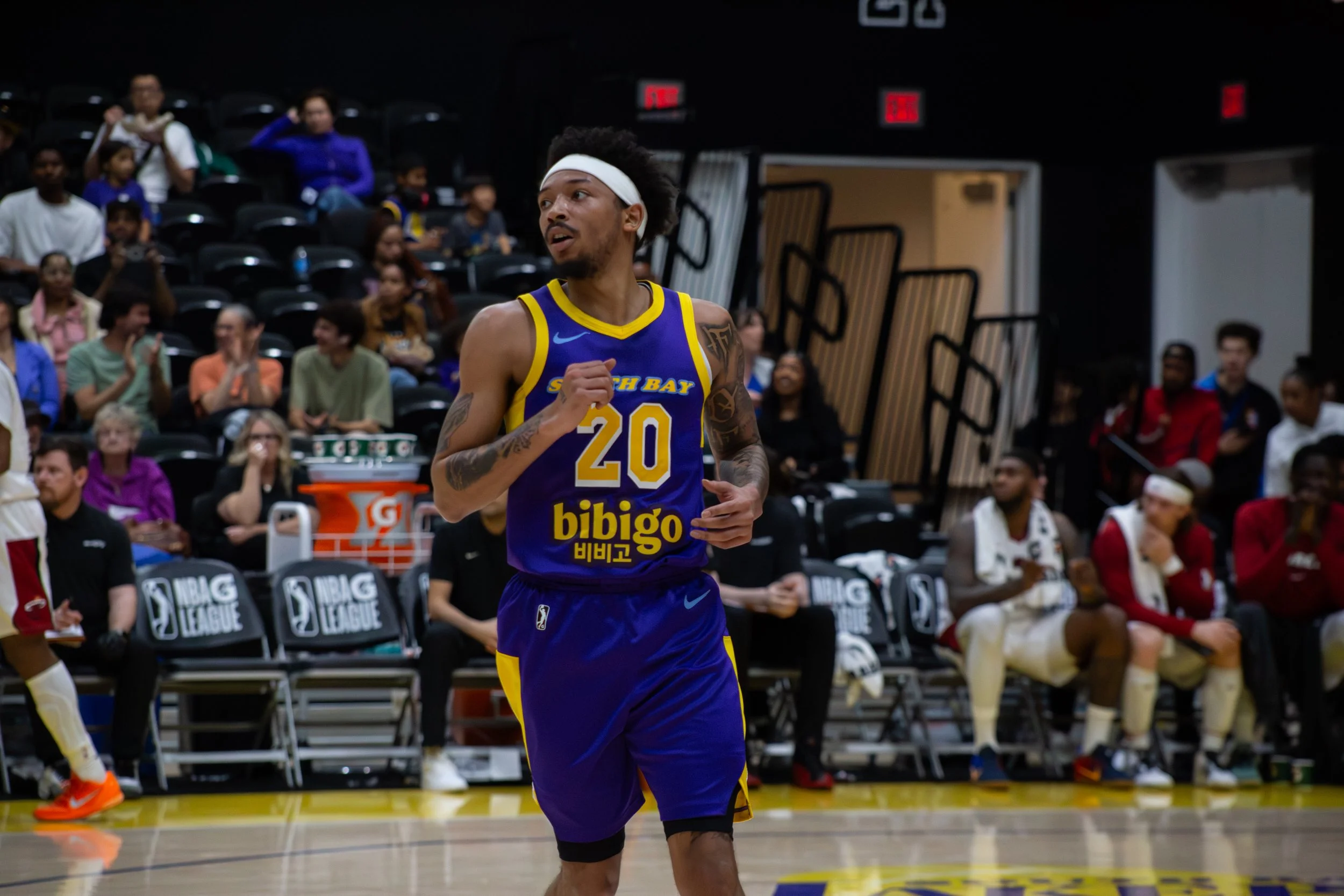 A basketball player wearing a blue and yellow jersey with the number 20, headband, and tattoos, running on the court during a game with spectators in the background.